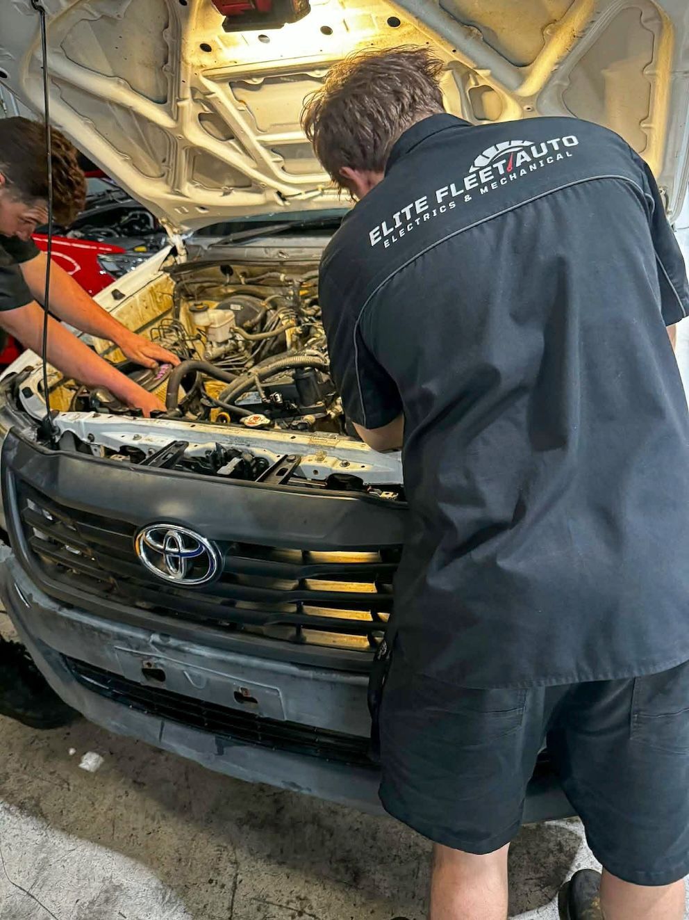 Two mechanics in black uniforms work on the open engine bay of a silver Toyota truck inside a garage— Elite Fleet Auto in Cannonvale, QLD