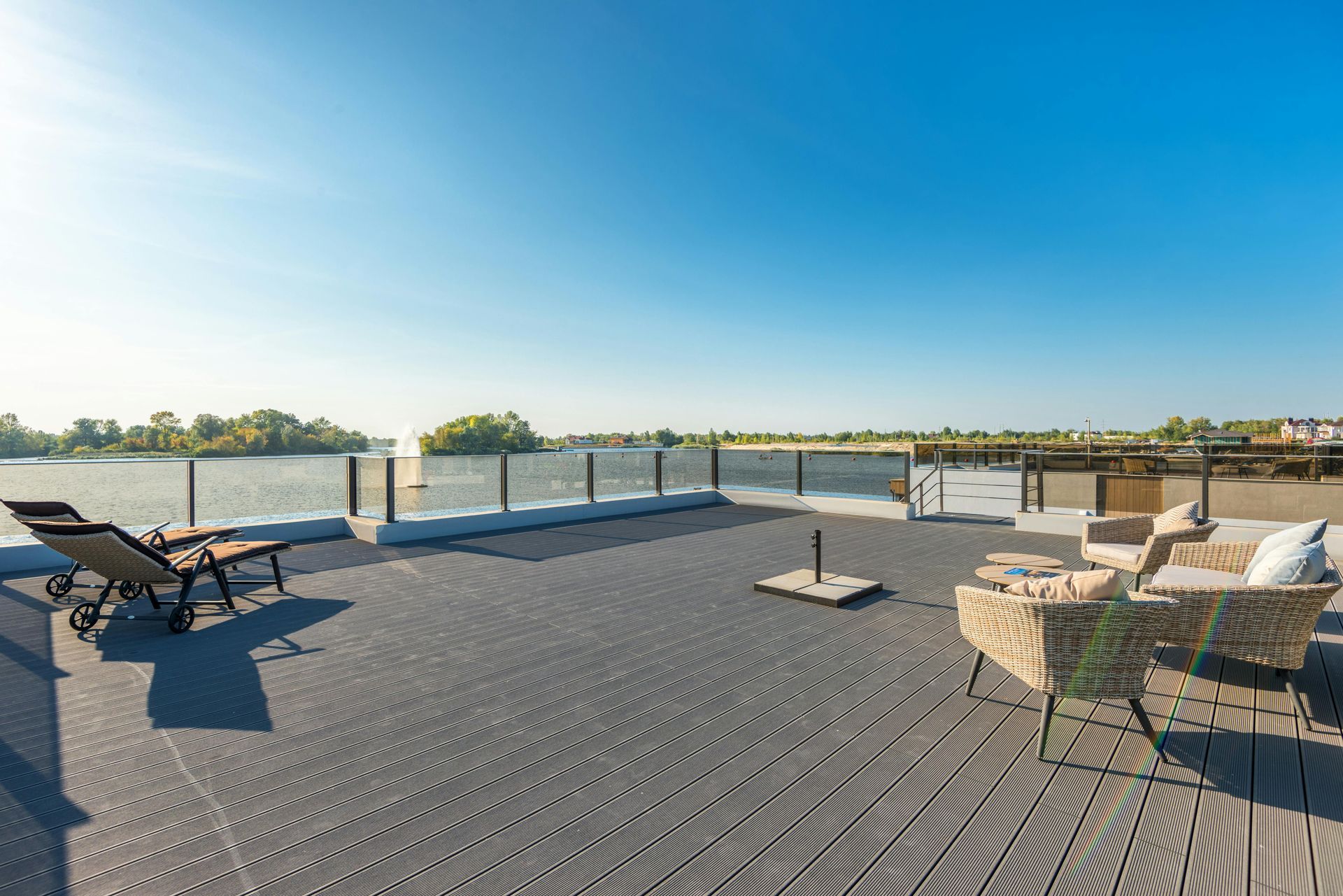 A sunny, gray-decked outdoor patio overlooking a calm lake, featuring wicker lounge chairs and a modern glass railing.