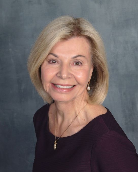 Smiling woman with blonde hair, wearing a purple top and pearl earrings, looking at the camera.