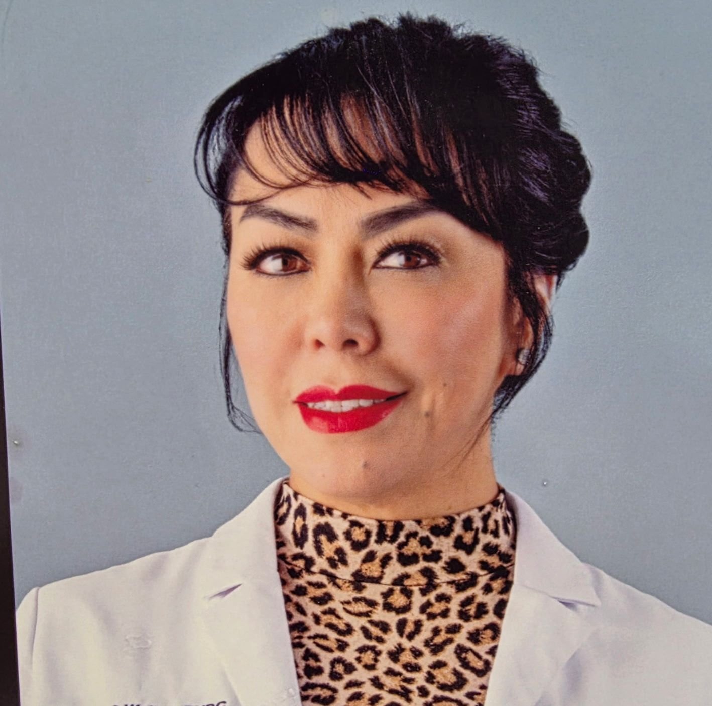 A person with dark hair styled with bangs wears a leopard-print top under a white lab coat, posing against a grey backdrop.