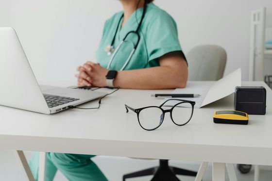 A person in light green scrubs and a stethoscope sits at a desk with a laptop, glasses, and office supplies.