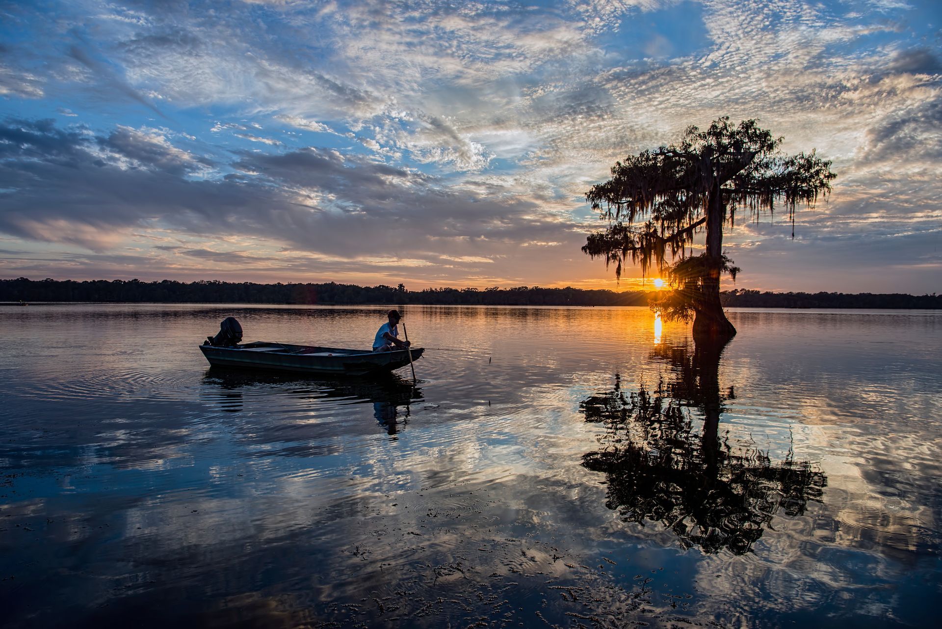 Man in boat fishing on calm lake at sunset, tree silhouetted, reflected in water, vibrant sky.
