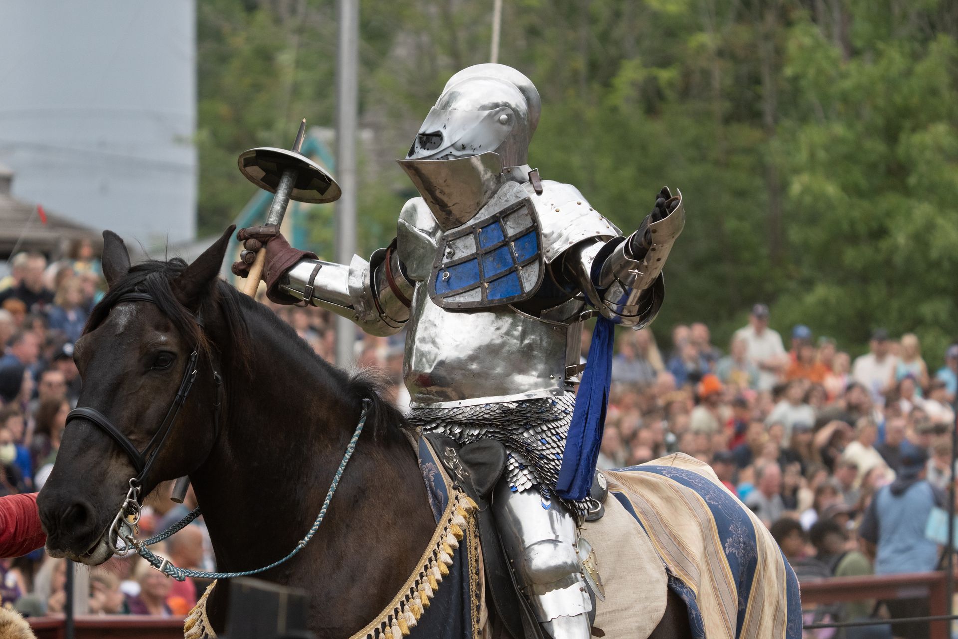 Knight in silver armor on horseback, holding a sword and gesturing, at a Renaissance fair.