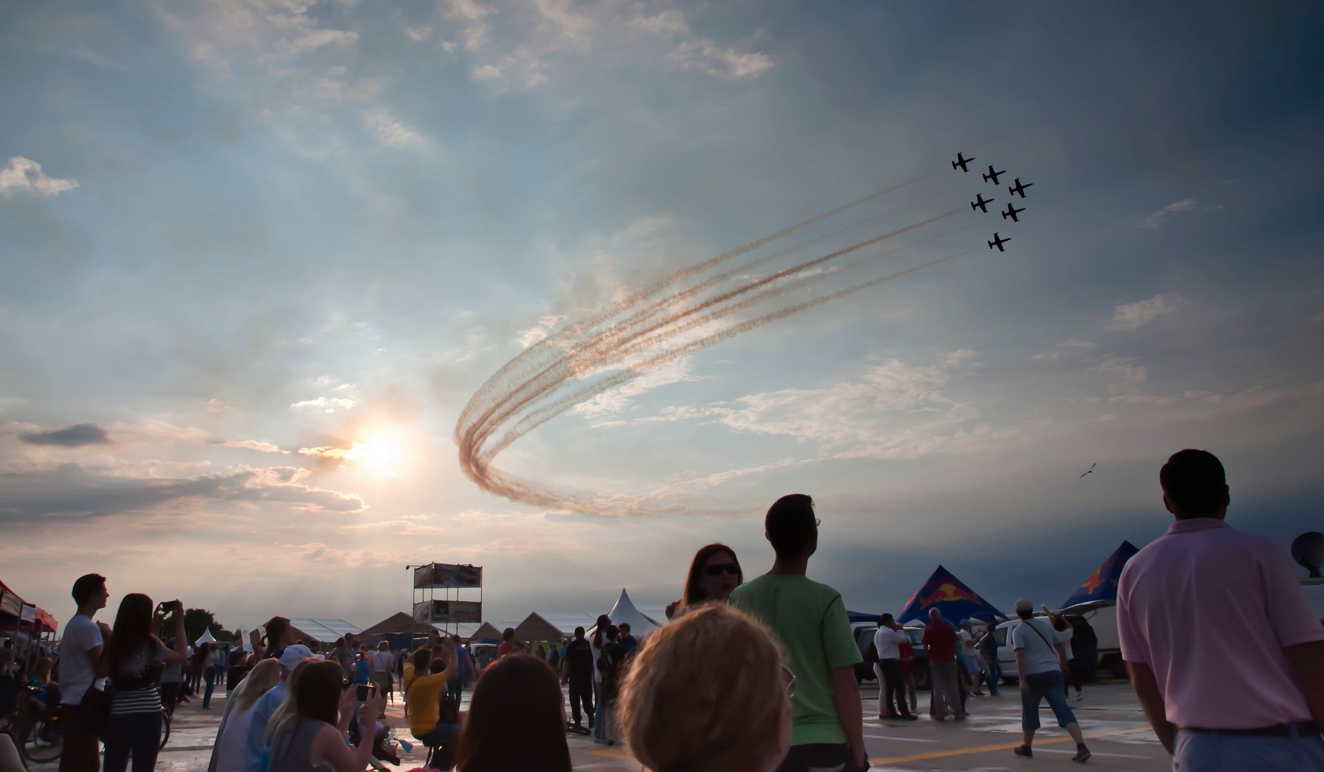 Airshow: Jets flying in formation, leaving red smoke trails, over a crowd at sunset.