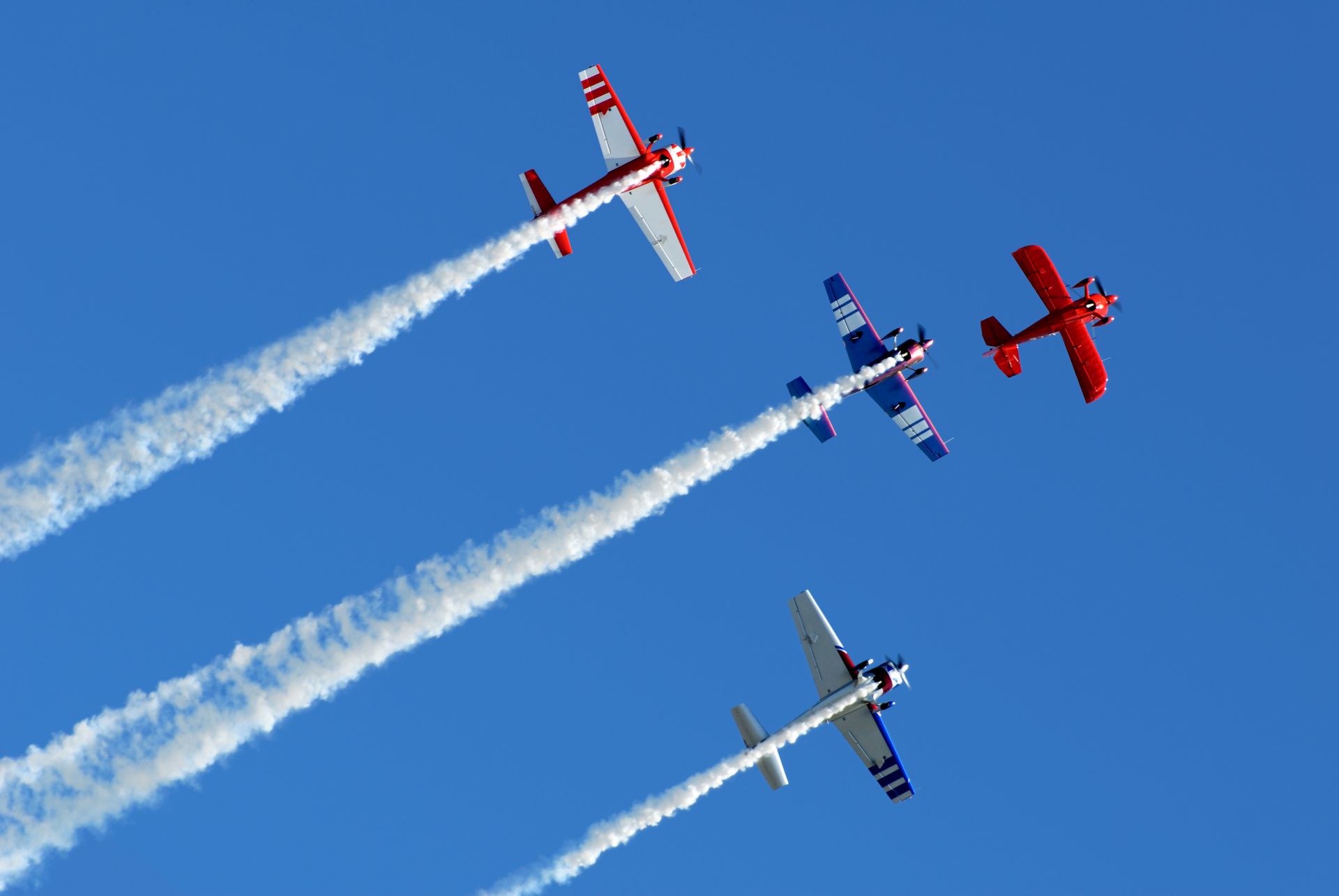Four biplanes in formation, emitting white smoke trails against a clear, blue sky.