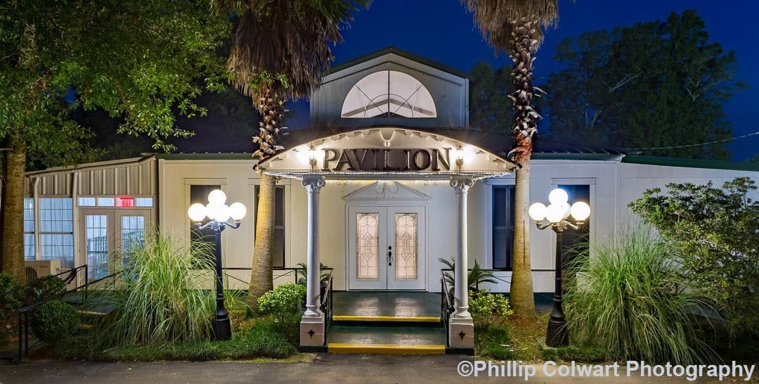 The pavilion is lit up at night with palm trees in front of it.
