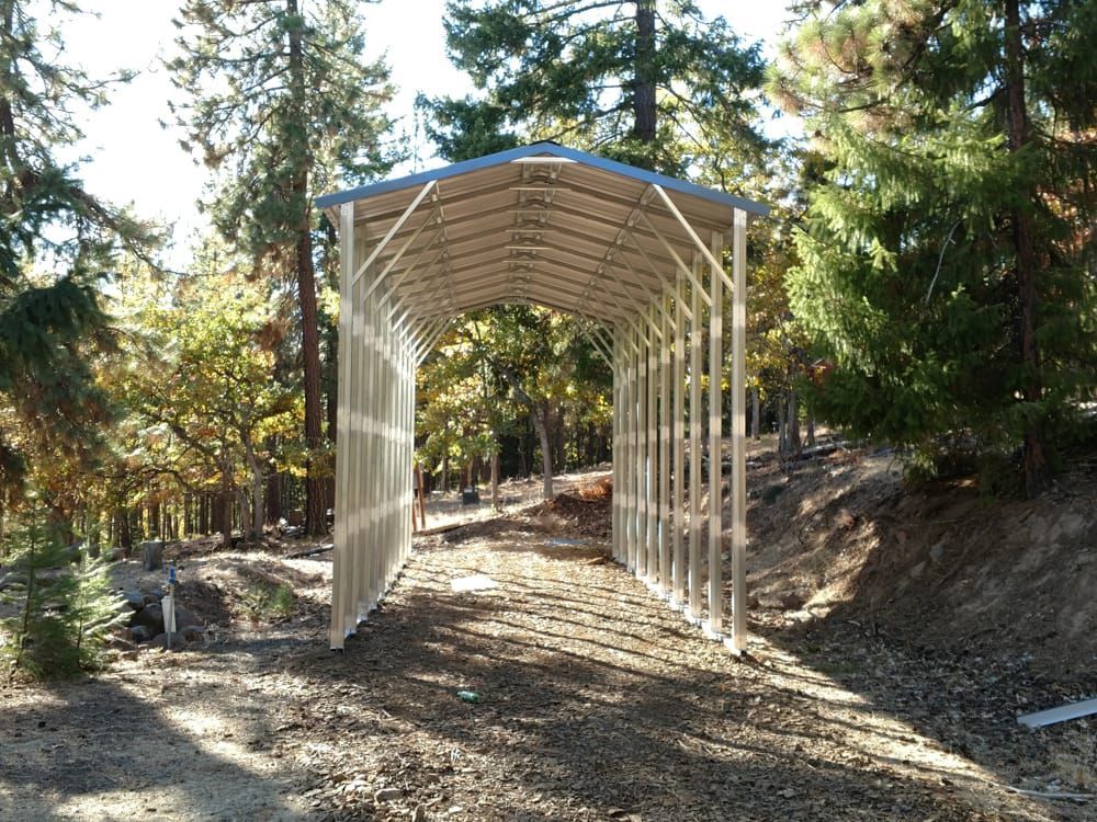 A metal carport is sitting on the side of a dirt road surrounded by trees.
