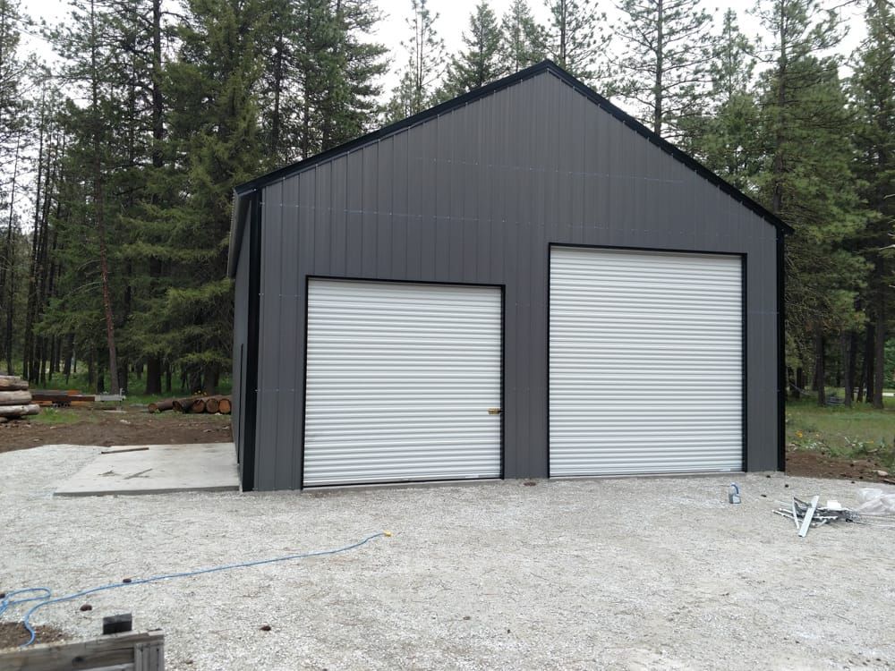 A black garage with two white garage doors is surrounded by trees