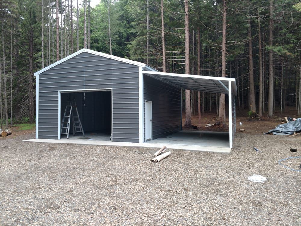 A metal garage with a carport attached to it is being built in the middle of a forest.