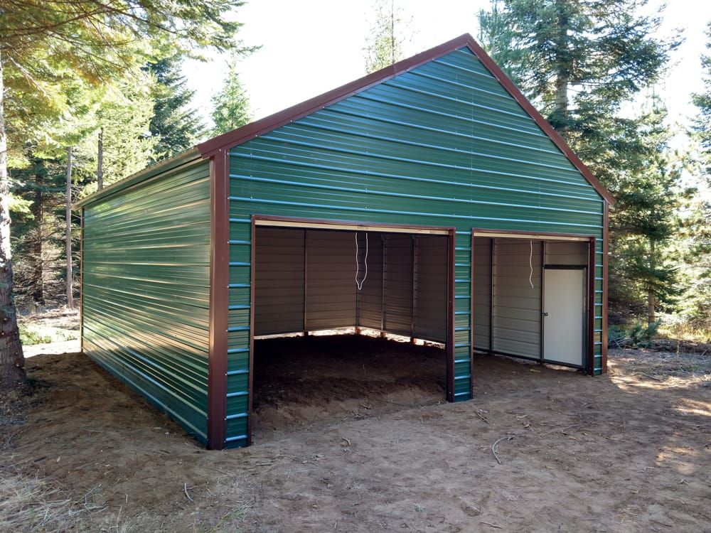 A green metal garage with a brown roof is sitting in the middle of a dirt field.