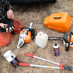 A man is kneeling down in the grass with a chainsaw and other tools.