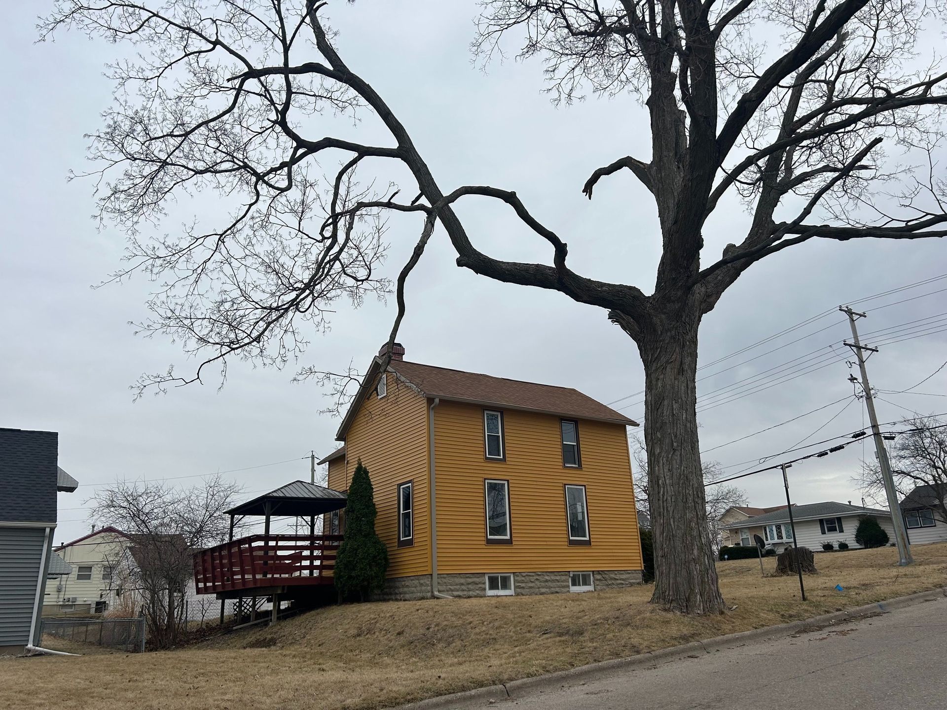 Yellow house with brown roof and red deck, next to a large bare tree on a cloudy day.