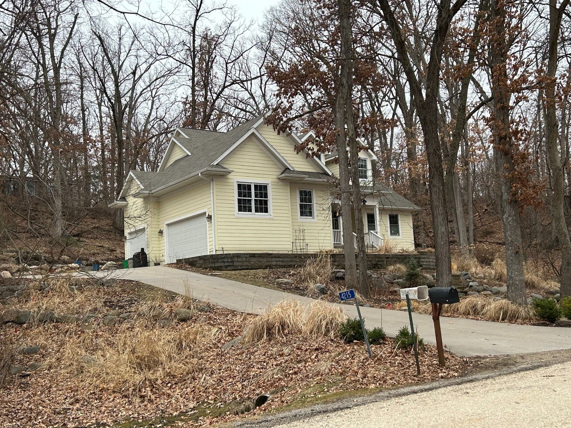 Yellow house with attached garage, driveway, and mailbox, surrounded by leafless trees.