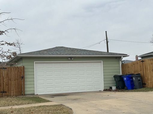 Green garage with white door, three trash cans, and a wooden fence on a cloudy day.