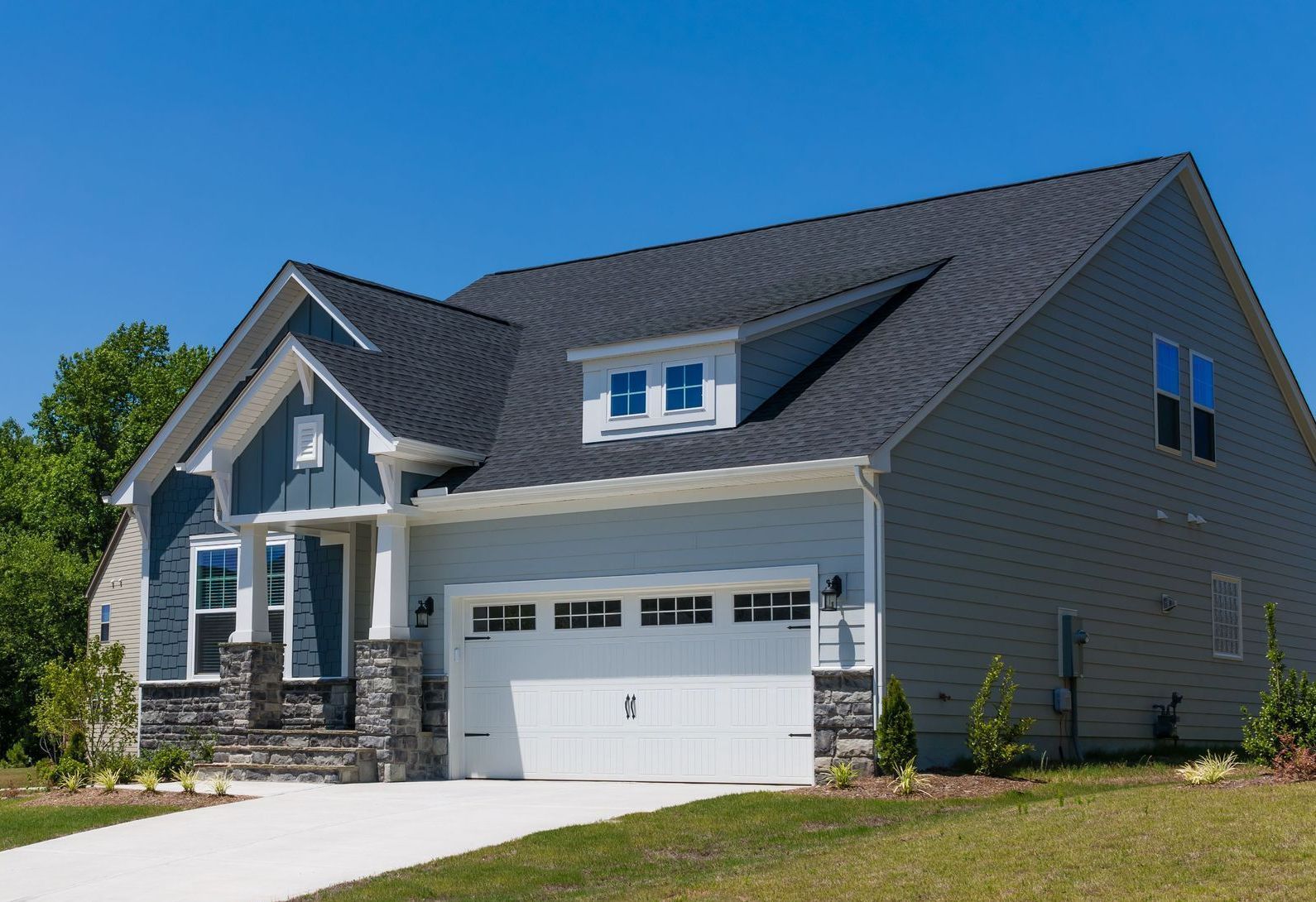 A light blue house with a white garage door, dark gray roof, and stone accents under a clear blue sky.