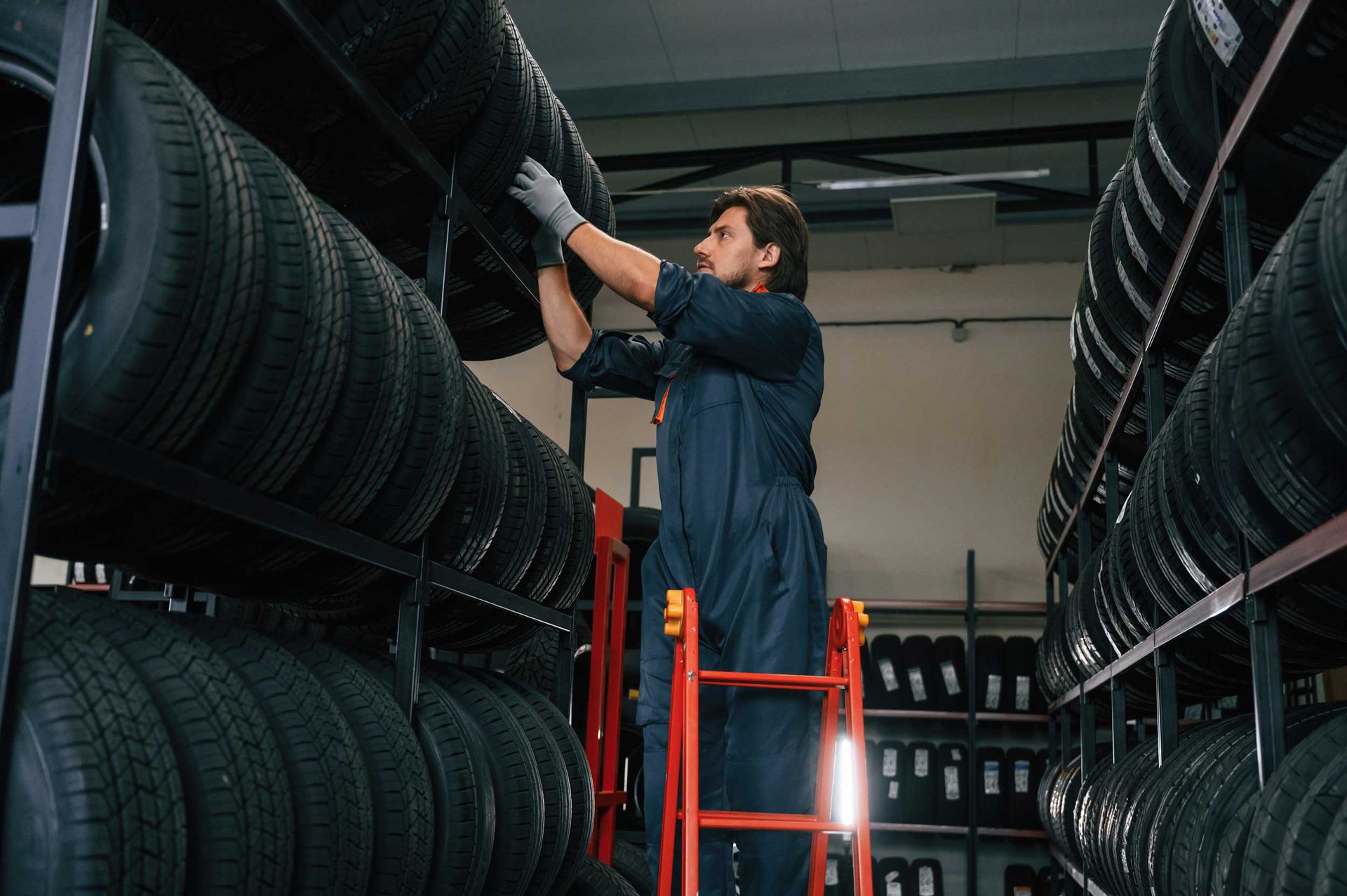 Person on ladder organizing tires in a warehouse with shelves full of stacked tires.