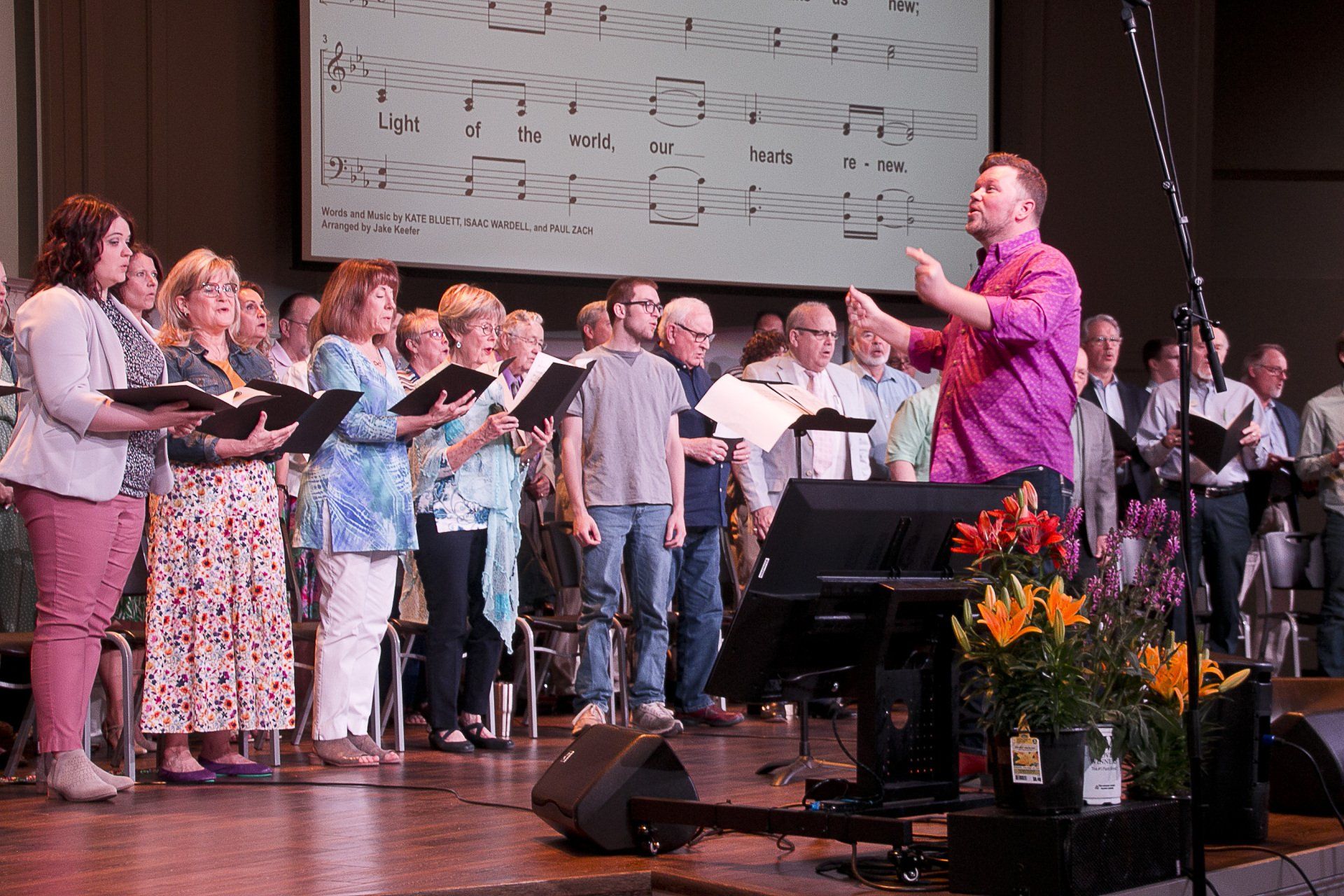 A man in a purple shirt is leading a choir on stage