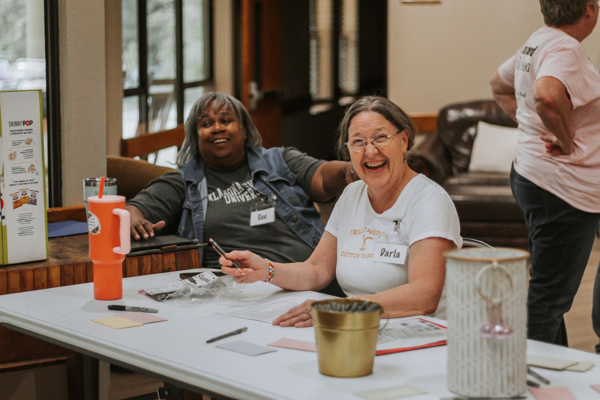 Two smiling women at a table with papers, pens, and a drink, in an indoor setting.