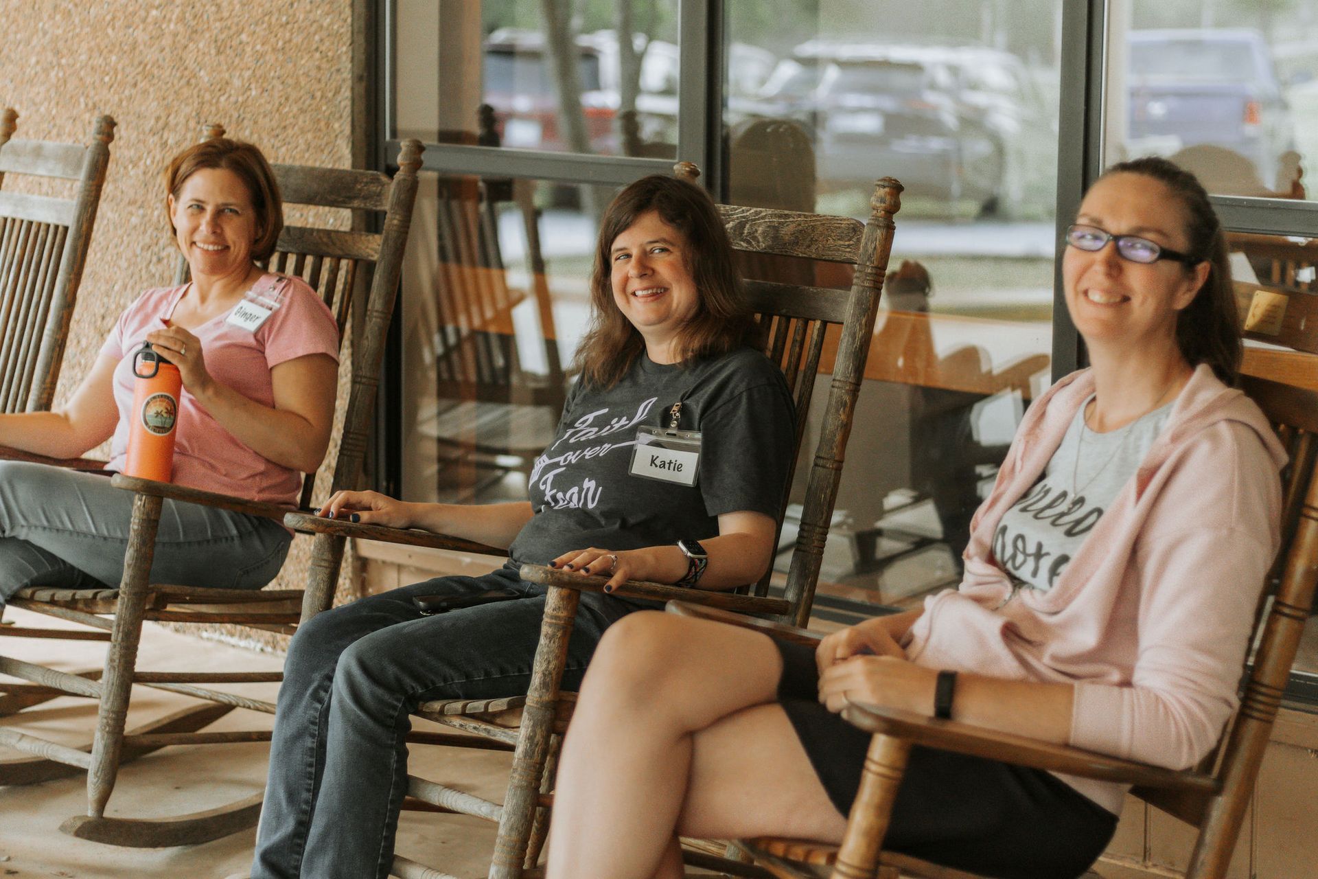 Three women smiling, sitting in rocking chairs on a porch. One holds an orange bottle. Sunny day.