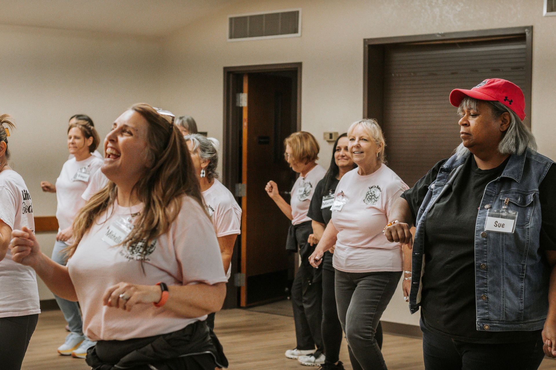 Group of women dancing in a room, some wearing pink shirts and denim.