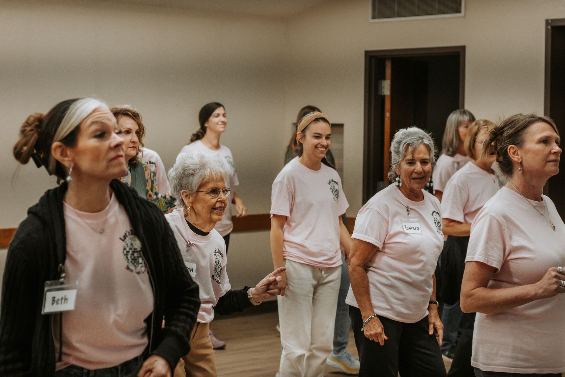 Women in pink shirts exercising in a light-filled room, smiling and engaged, possibly a fitness class.