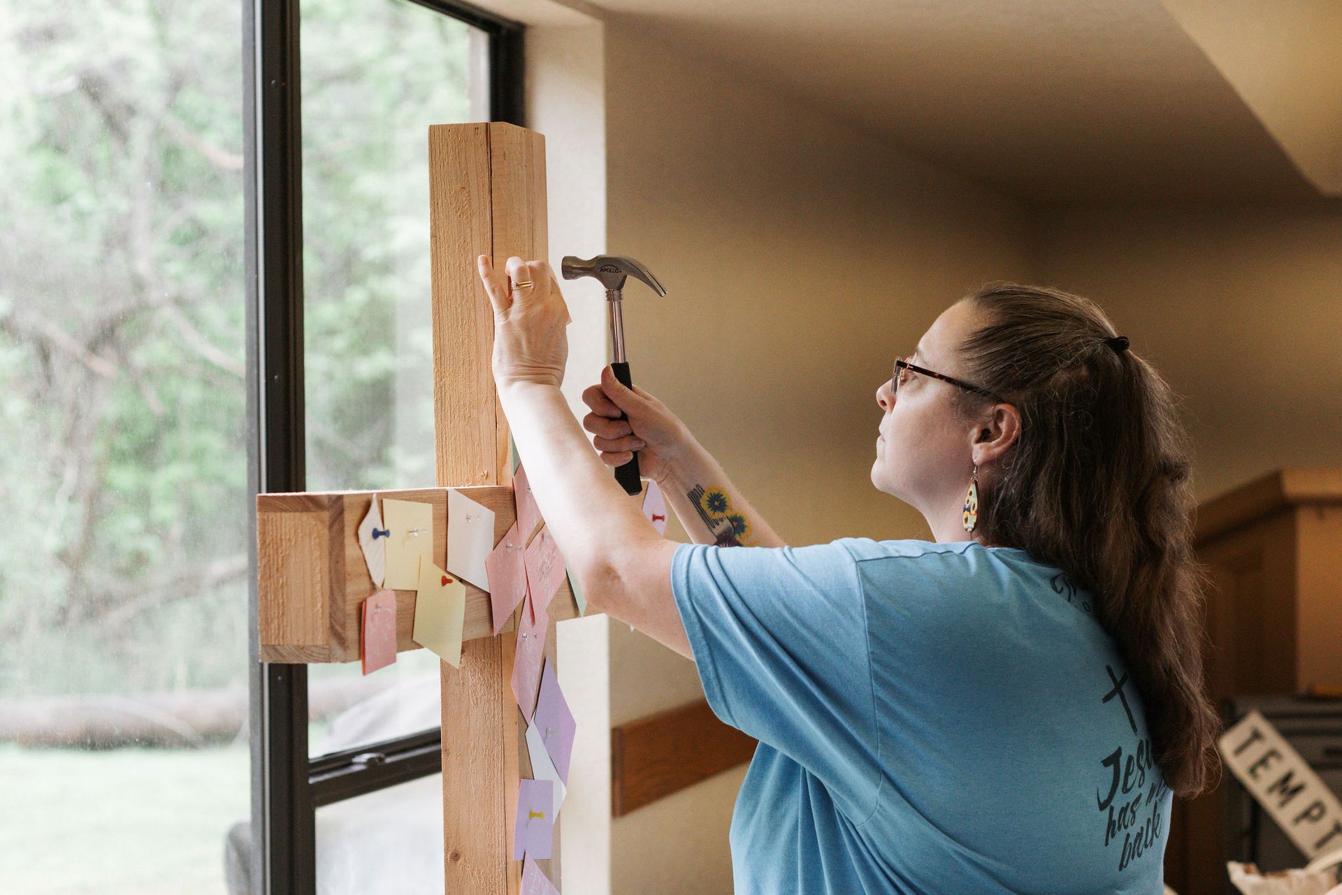 Woman hammering paper to a wooden cross by a window.
