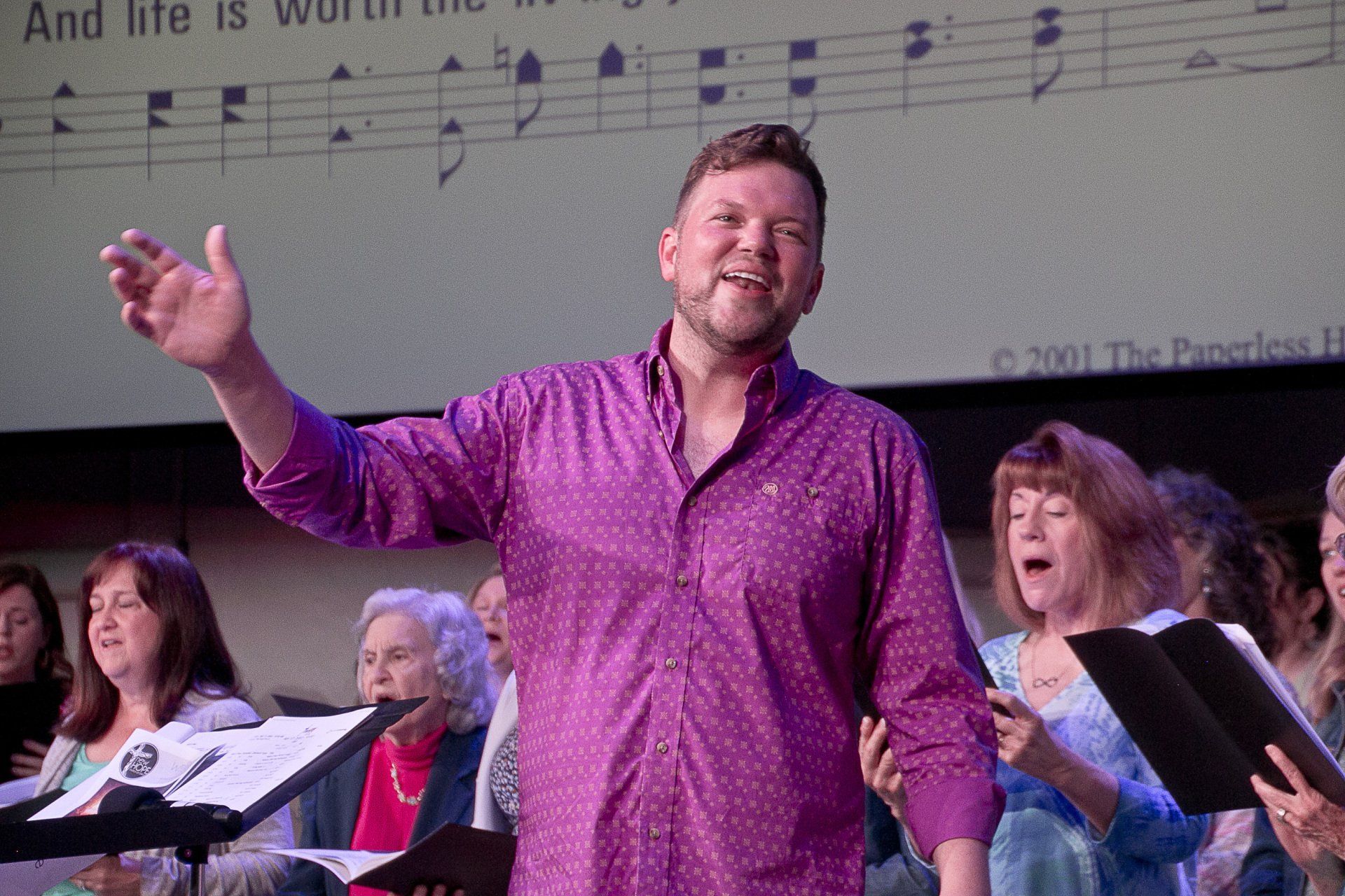 A man in a purple shirt is singing in front of a choir.