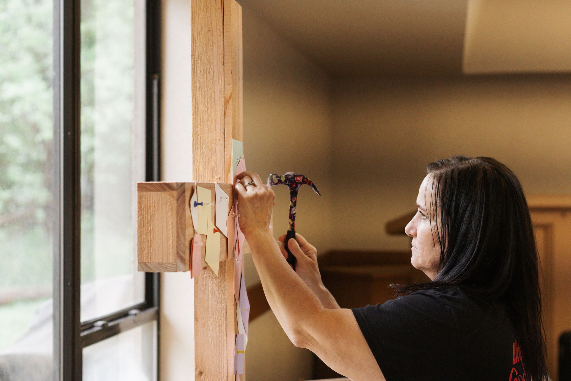 Woman hammering a note onto a wooden cross-shaped bulletin board near a window.