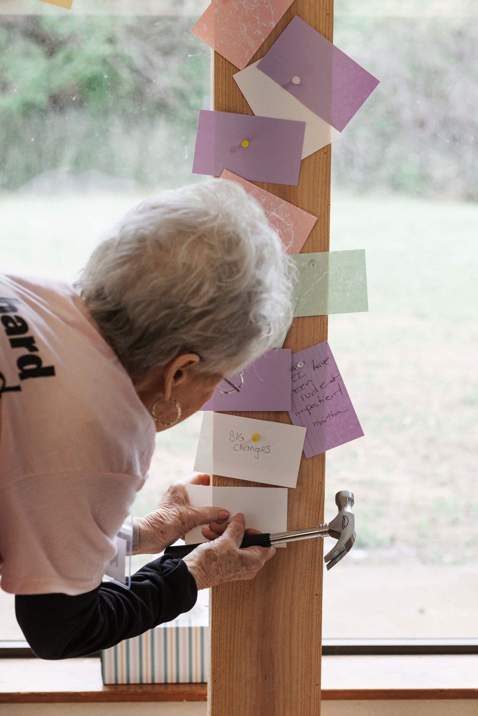 An elderly person nails cards to a wooden post, indoors near a window. They are wearing a pink shirt.
