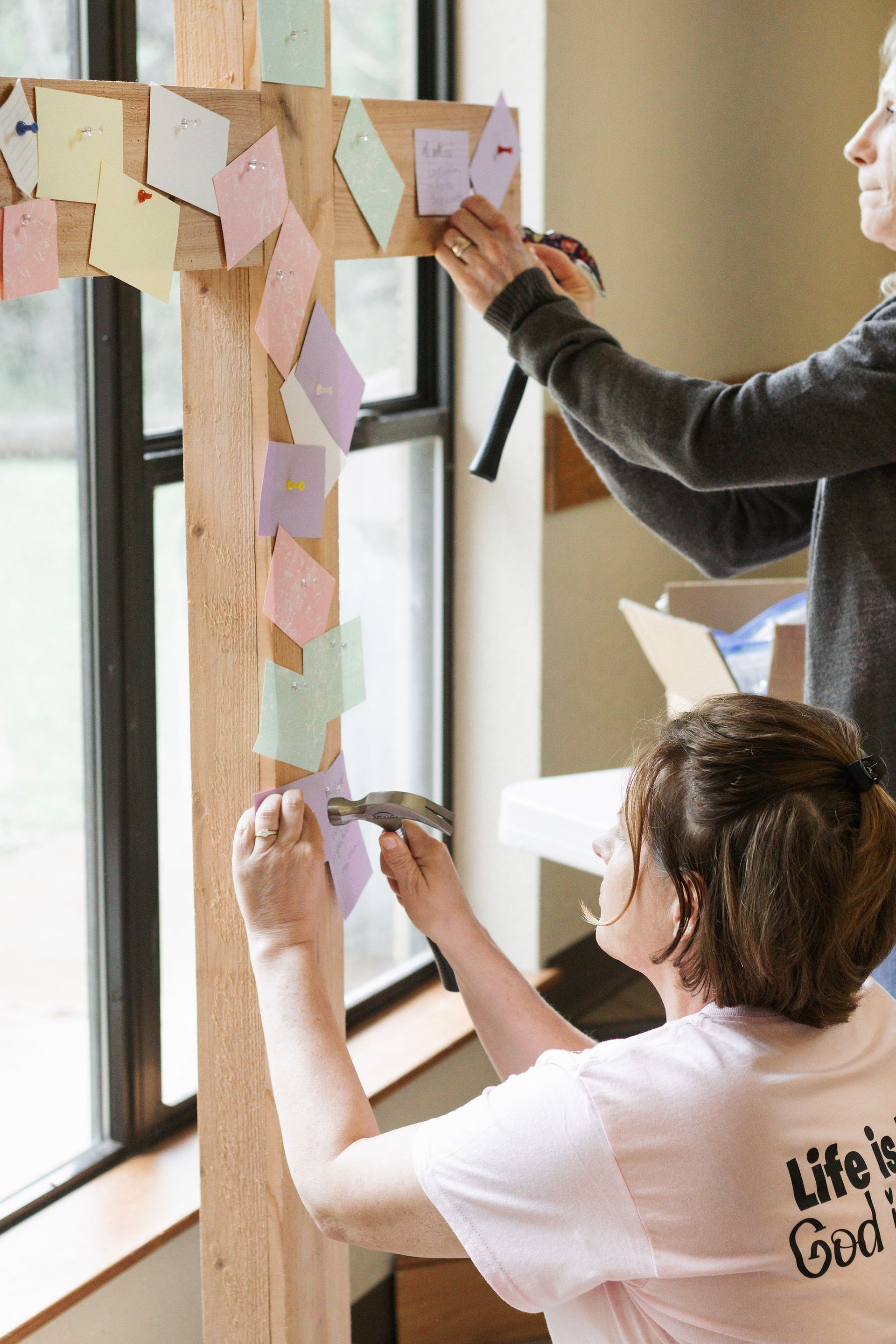 Two women attaching colorful notes to a wooden cross near a window.