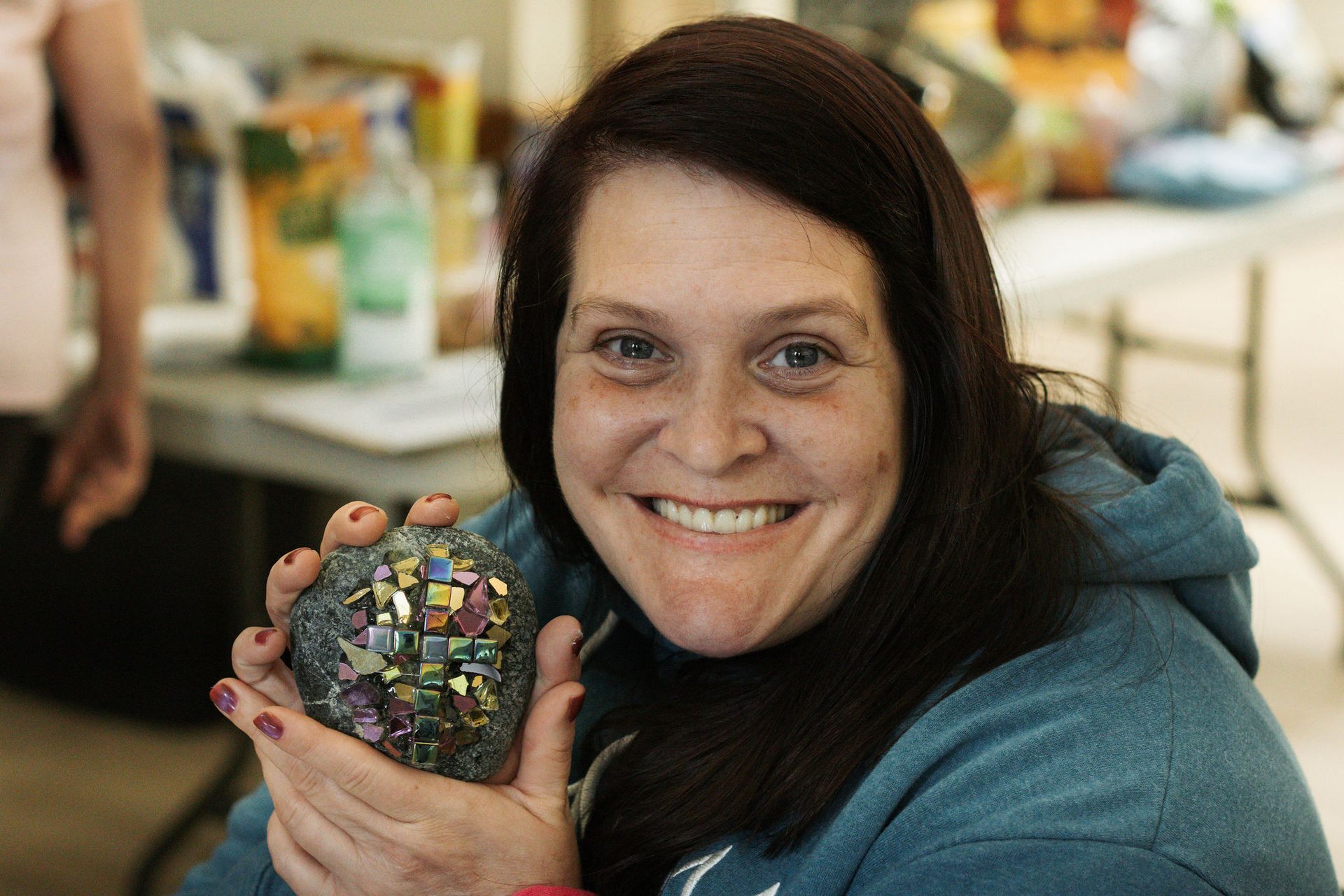 Woman smiles, holding a colorful mosaic-covered rock. She wears a blue hoodie in a bright room.
