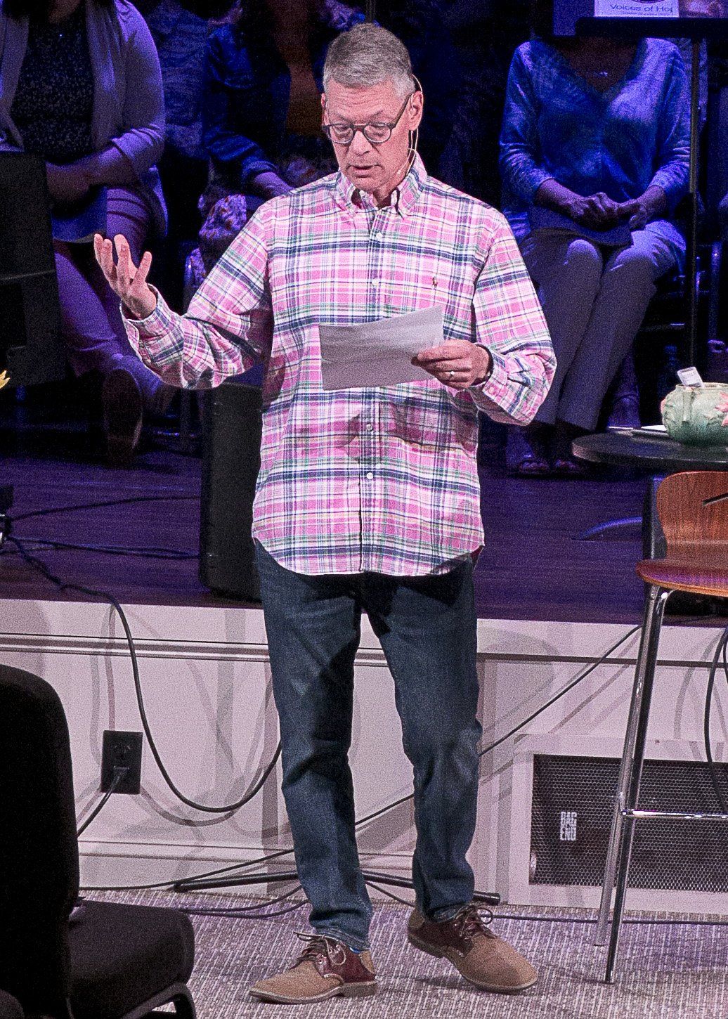 A man in a plaid shirt is standing on a stage holding a piece of paper.