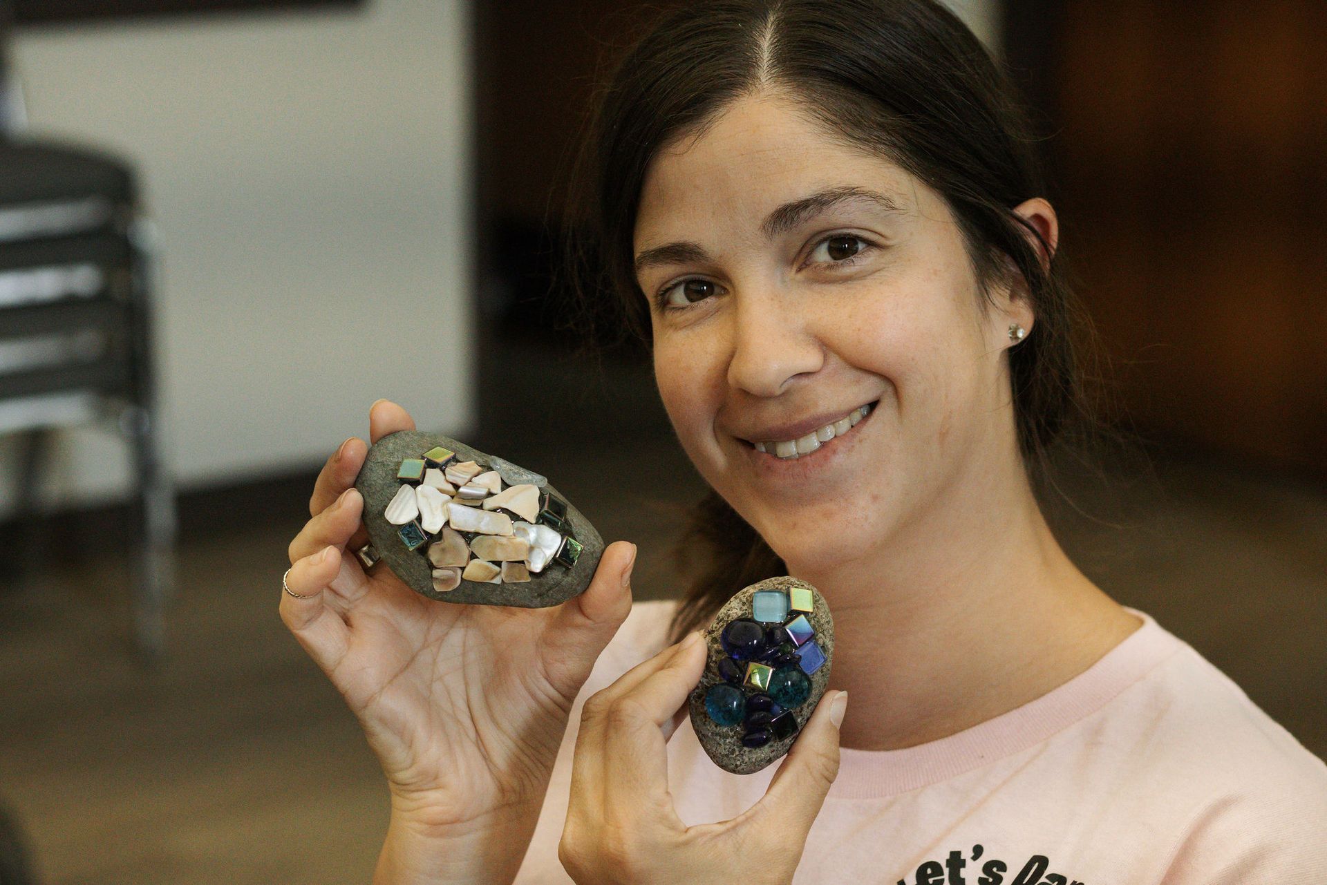Woman smiling, holding decorated rocks. Pale pink shirt, indoor setting.