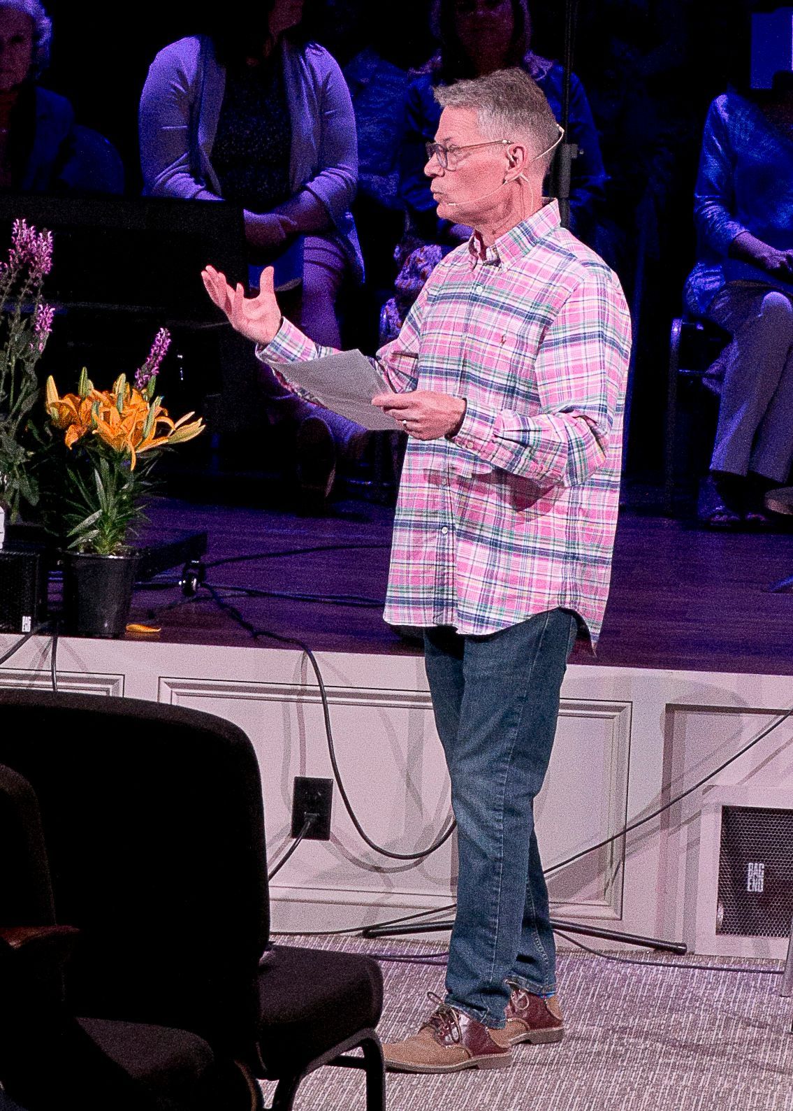 A man in a plaid shirt is standing on a stage holding a clipboard.