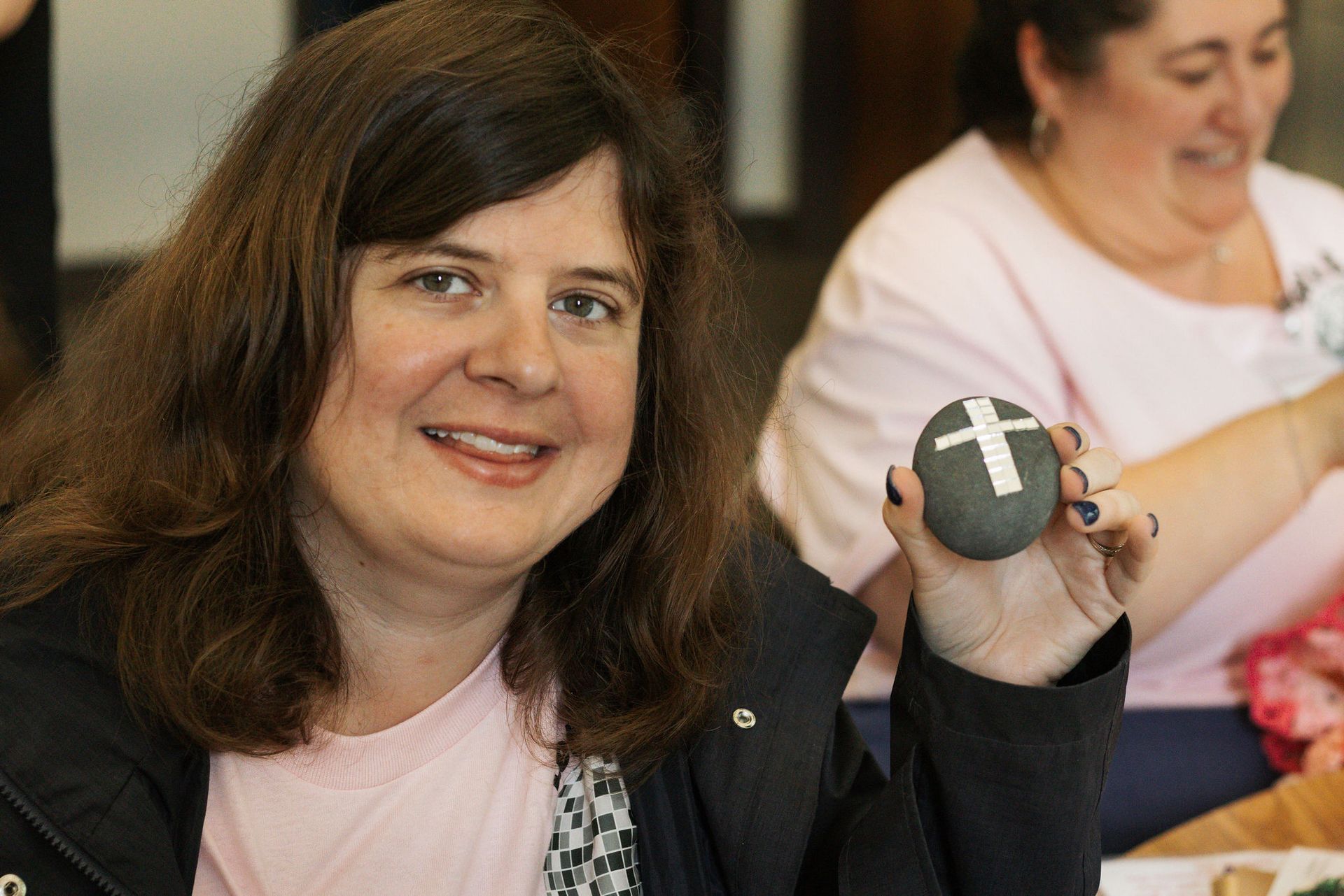 Woman smiling, holding a gray stone with a white cross painted on it. Pink shirt, dark jacket, indoor setting.