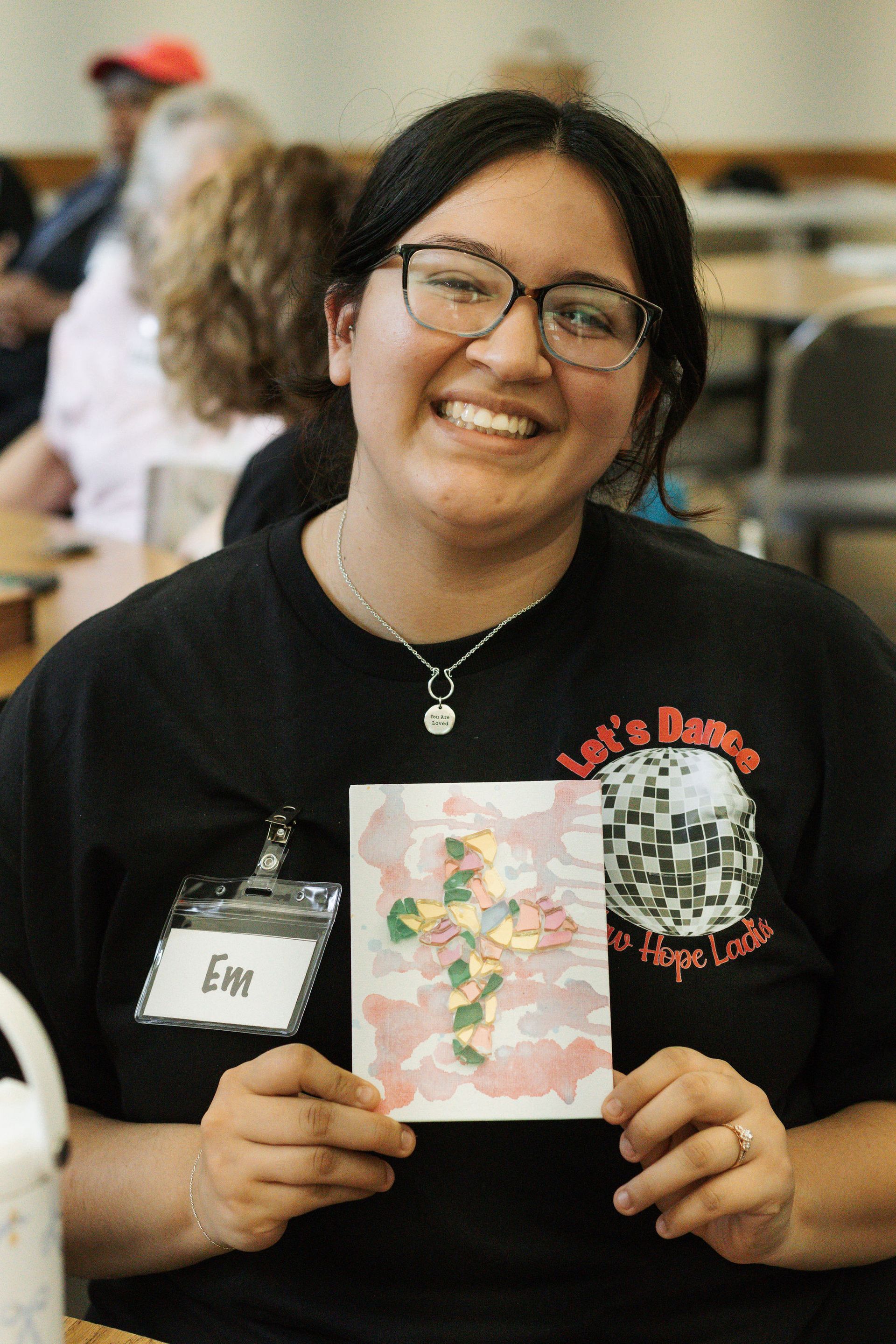 Smiling woman holds up a watercolor painting of a cross in a room.