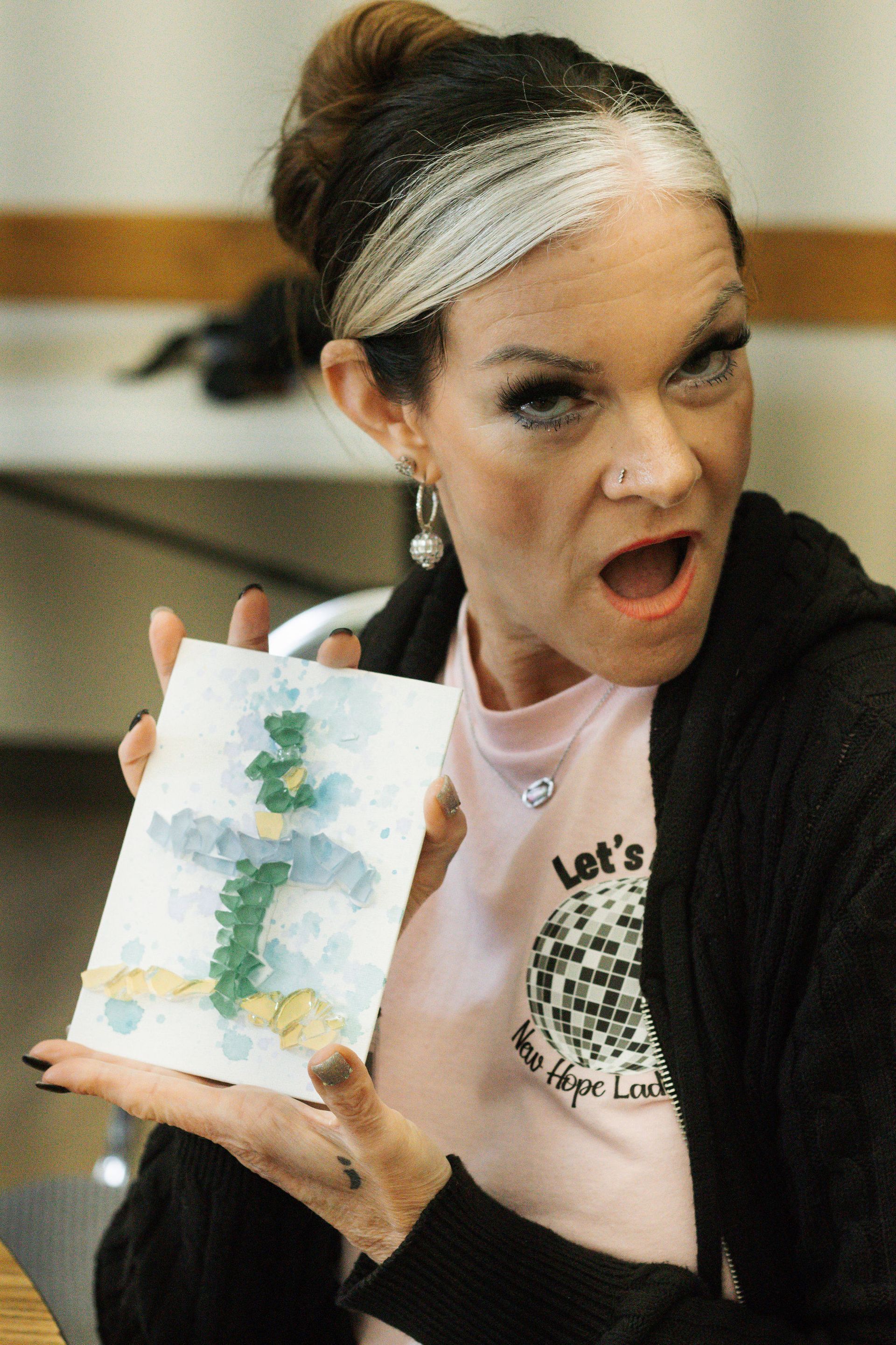 Woman with highlighted hair, holding up a watercolor painting, surprised expression, wearing a pink shirt.