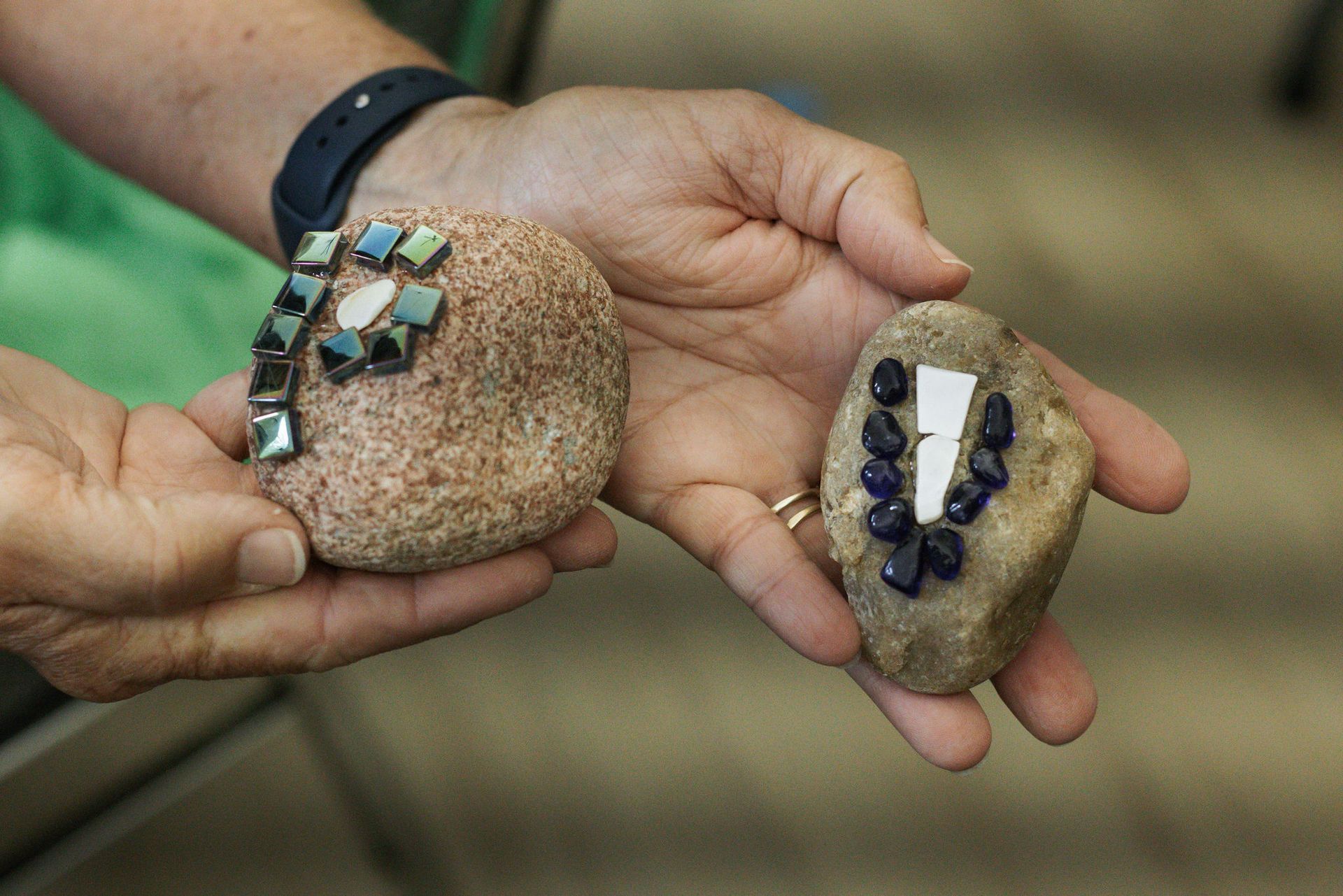 Person holding two decorated rocks, one with mosaic tiles, the other with blue stones.