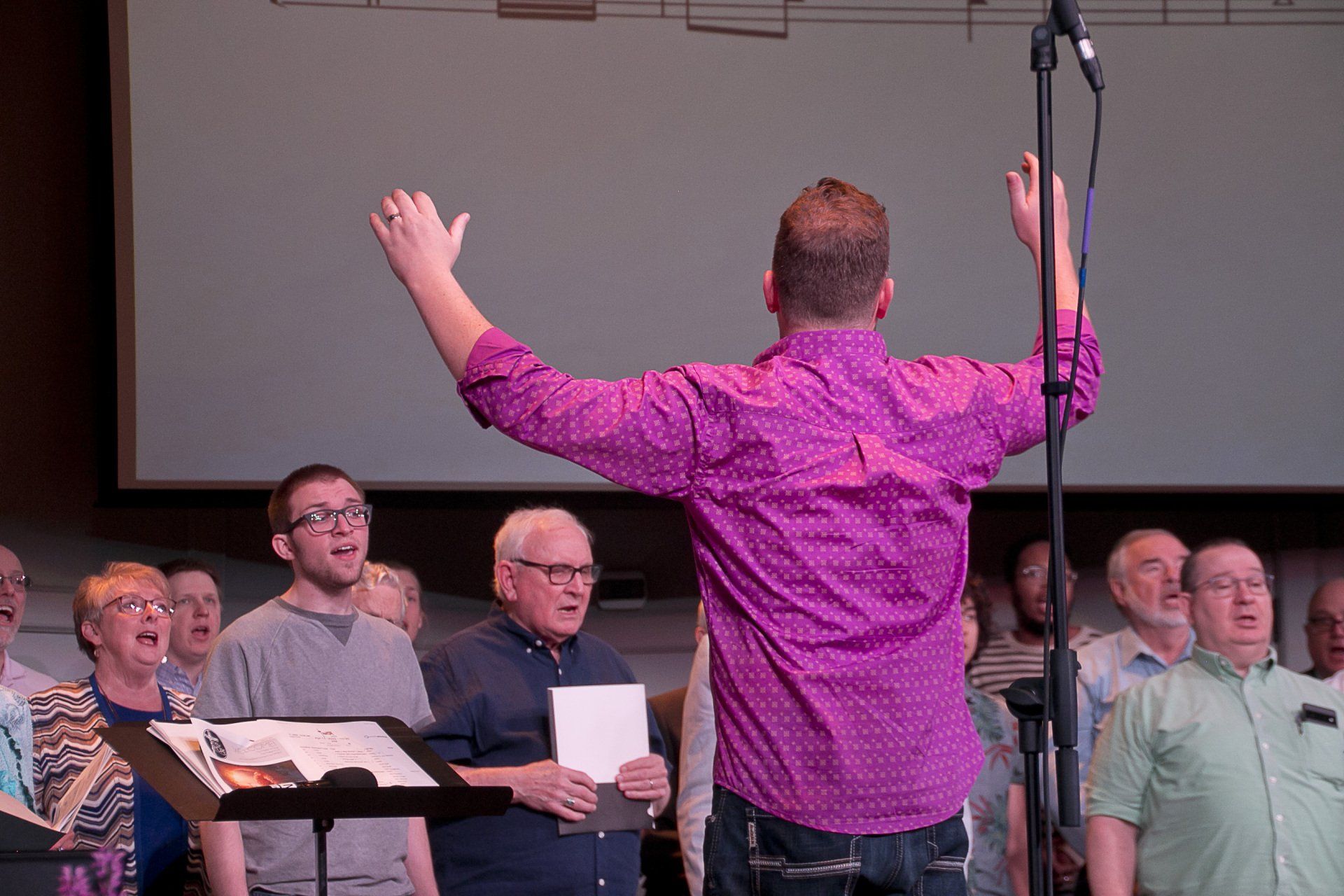 A man in a purple shirt is leading a choir on stage.