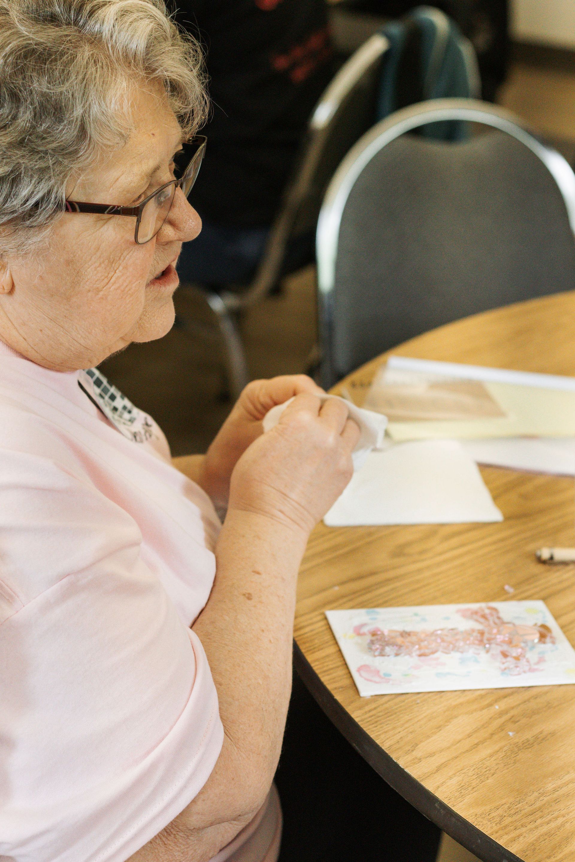 Woman with glasses crafting at a table; light pink shirt.