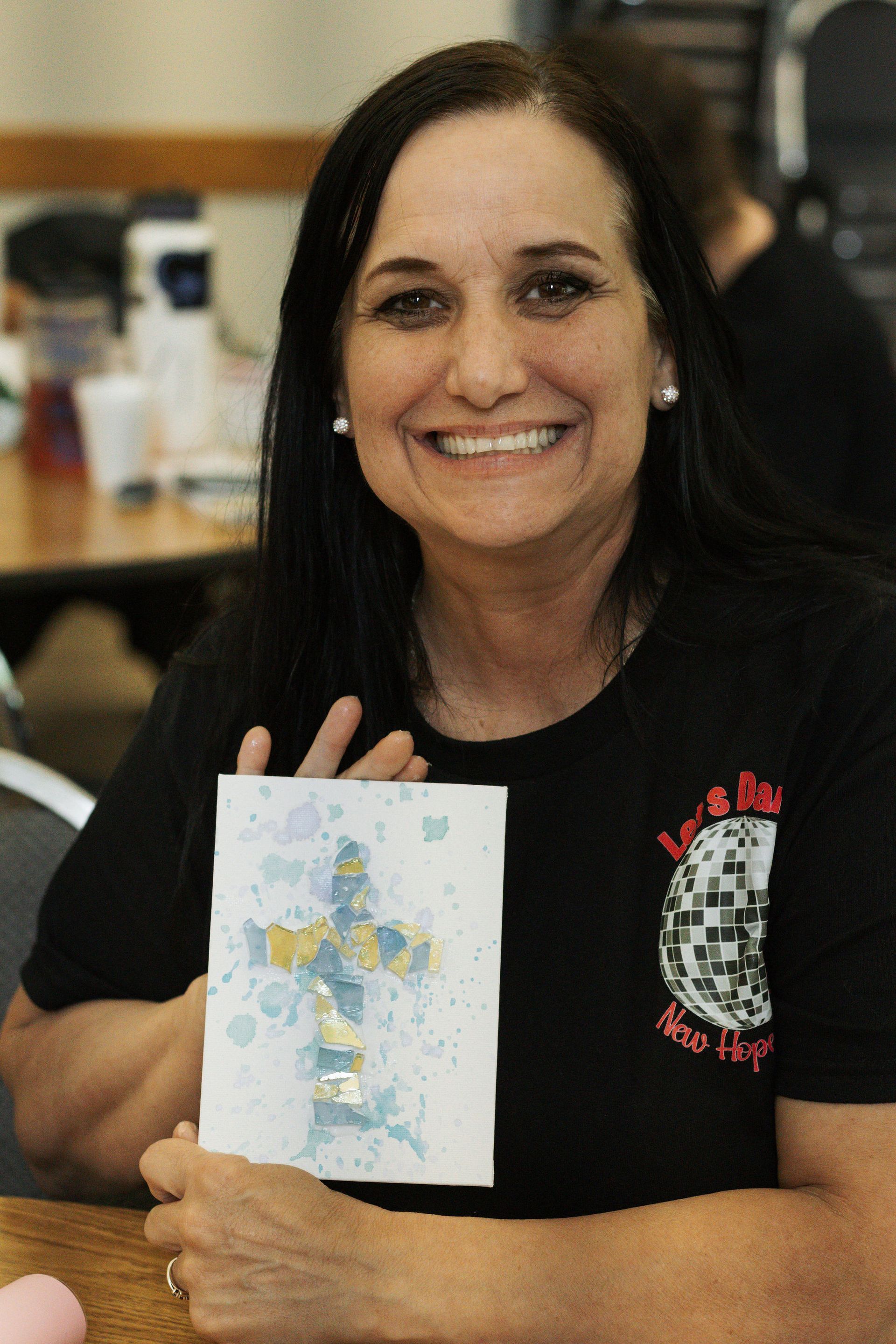 Woman holding a handmade card with a cross design, smiling in a room.