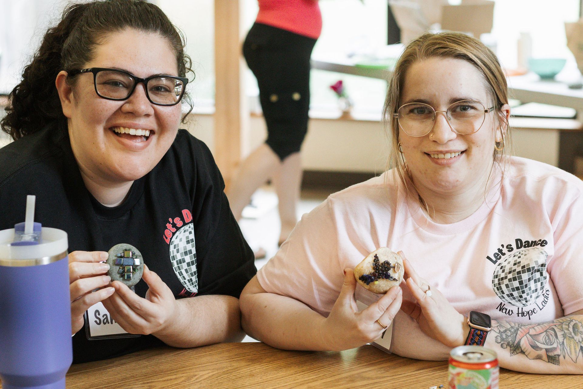 Two women smiling, holding decorated eggs and a muffin, indoors at a table.