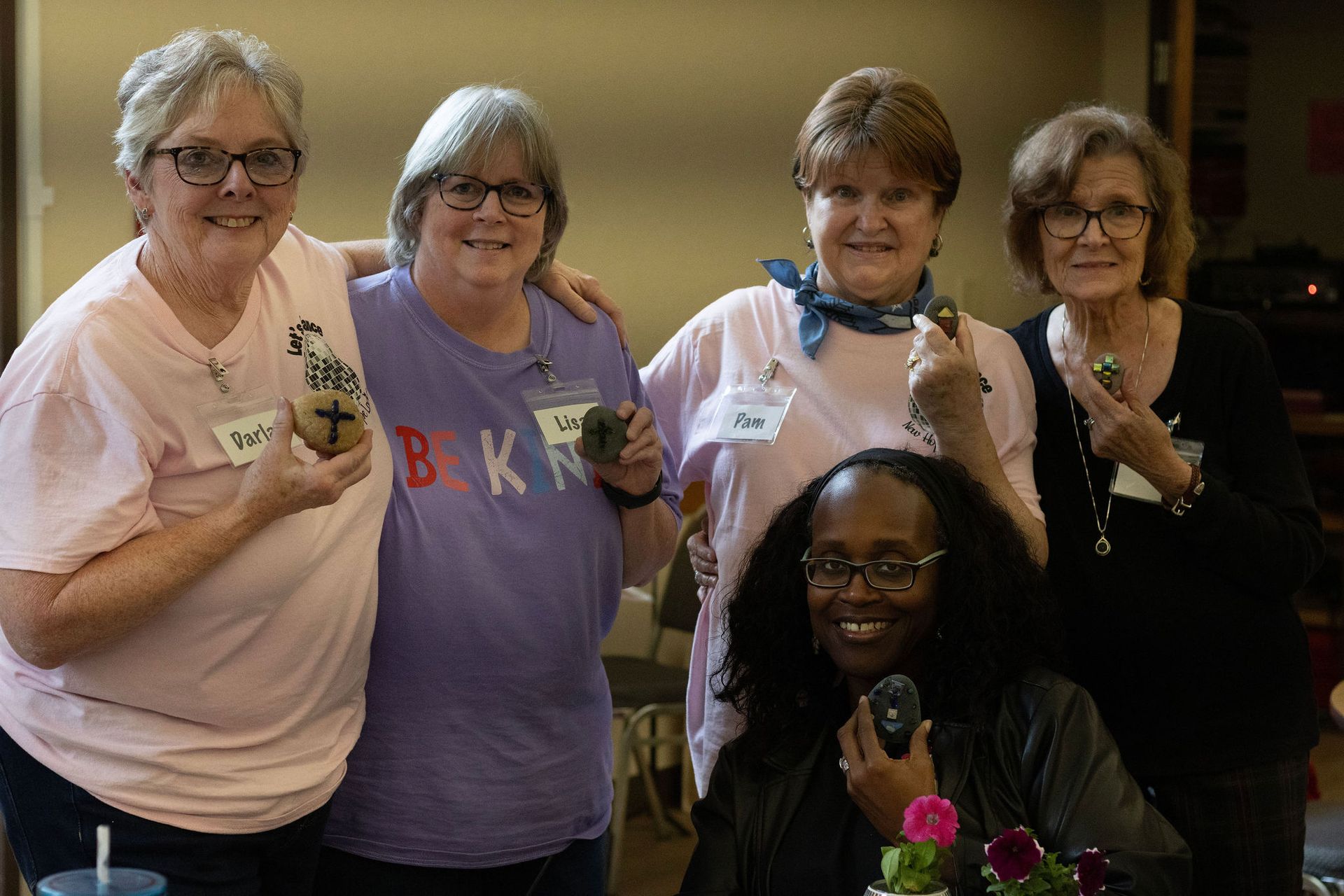 Five women smiling, posing together indoors, holding small objects. Pink and purple shirts, glasses.