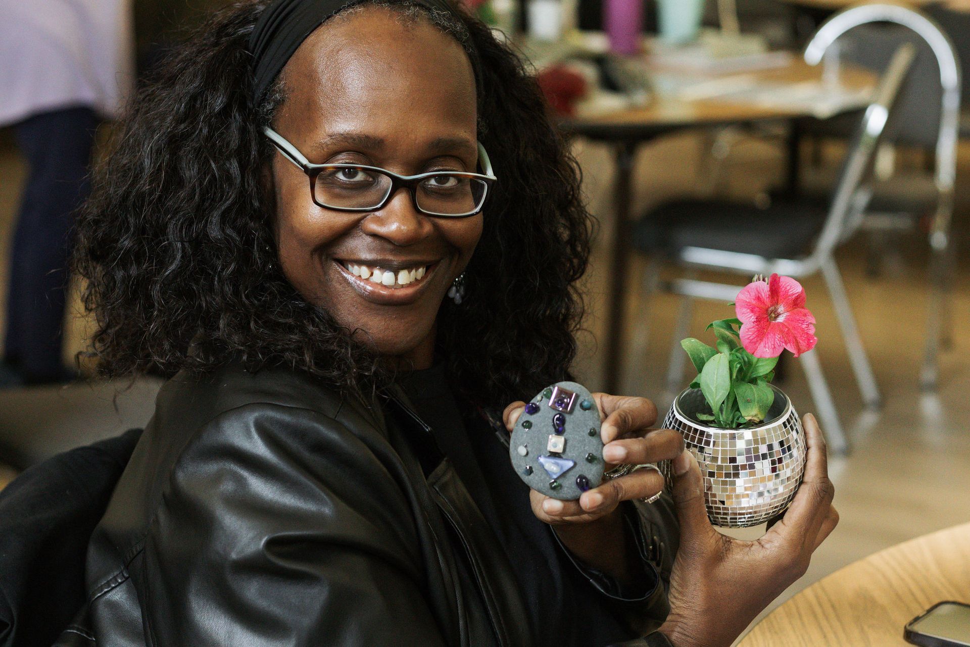 Woman with curly hair and glasses smiling, holding a small plant with pink flower and gray decorative object.