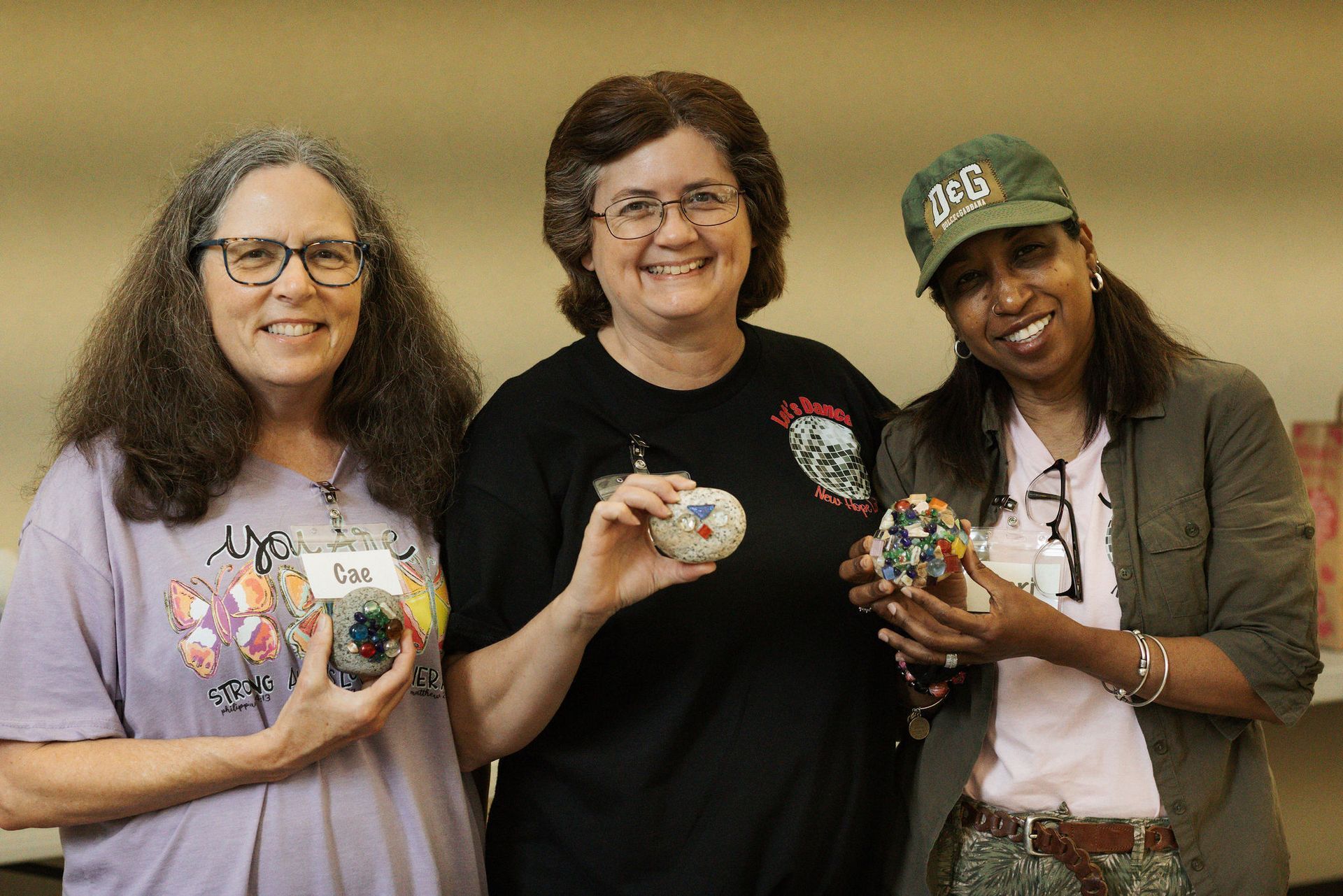 Three women smiling, holding decorated rocks. One wears glasses, another a black t-shirt, and the third a cap and jacket.