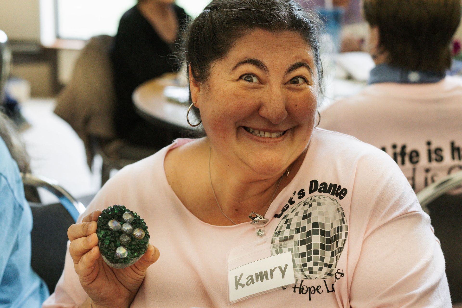Woman smiles, holding a dark green rock with lighter stones, wearing a pink shirt, in a room.