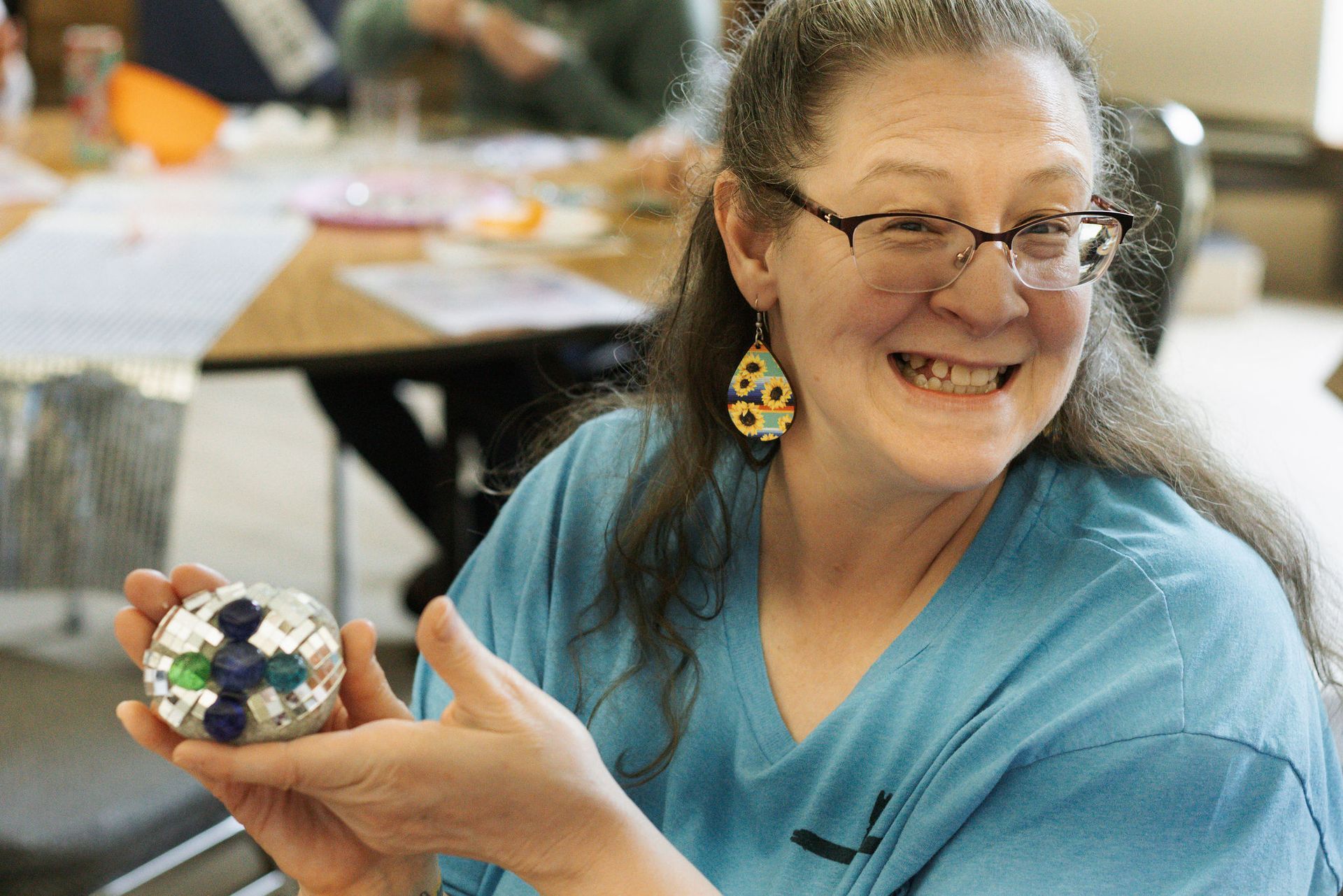 Woman with glasses smiling, holding mosaic artwork; indoors, wearing a blue shirt and sunflower earrings.