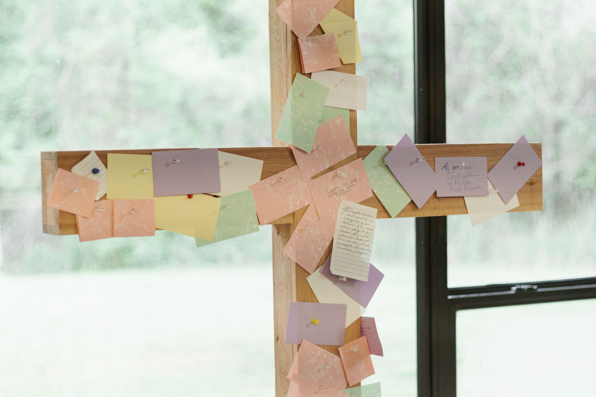 Wooden cross covered in colorful sticky notes, near a window.