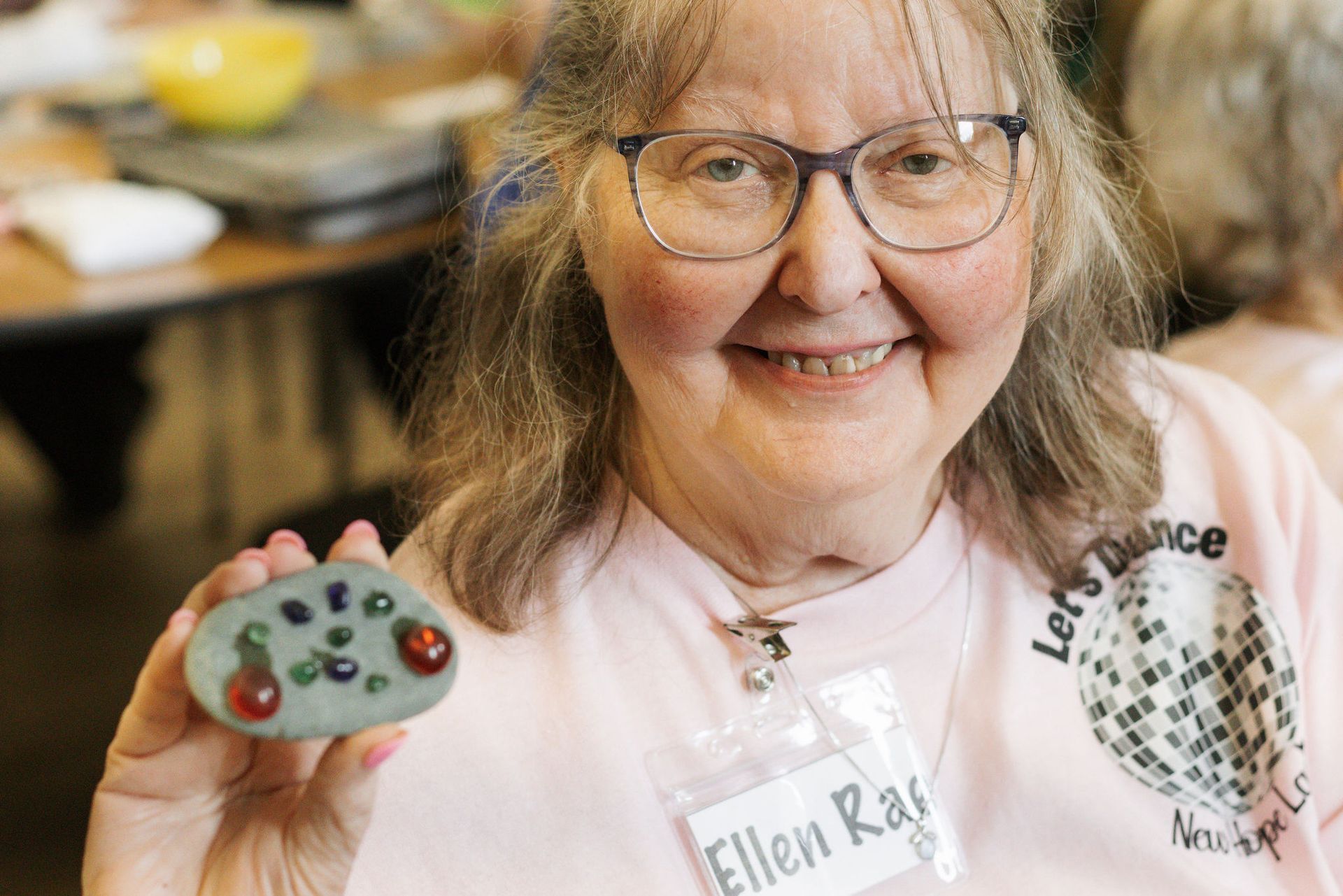 Smiling senior woman holding a painted rock resembling a video game controller. She wears glasses and a pink t-shirt.