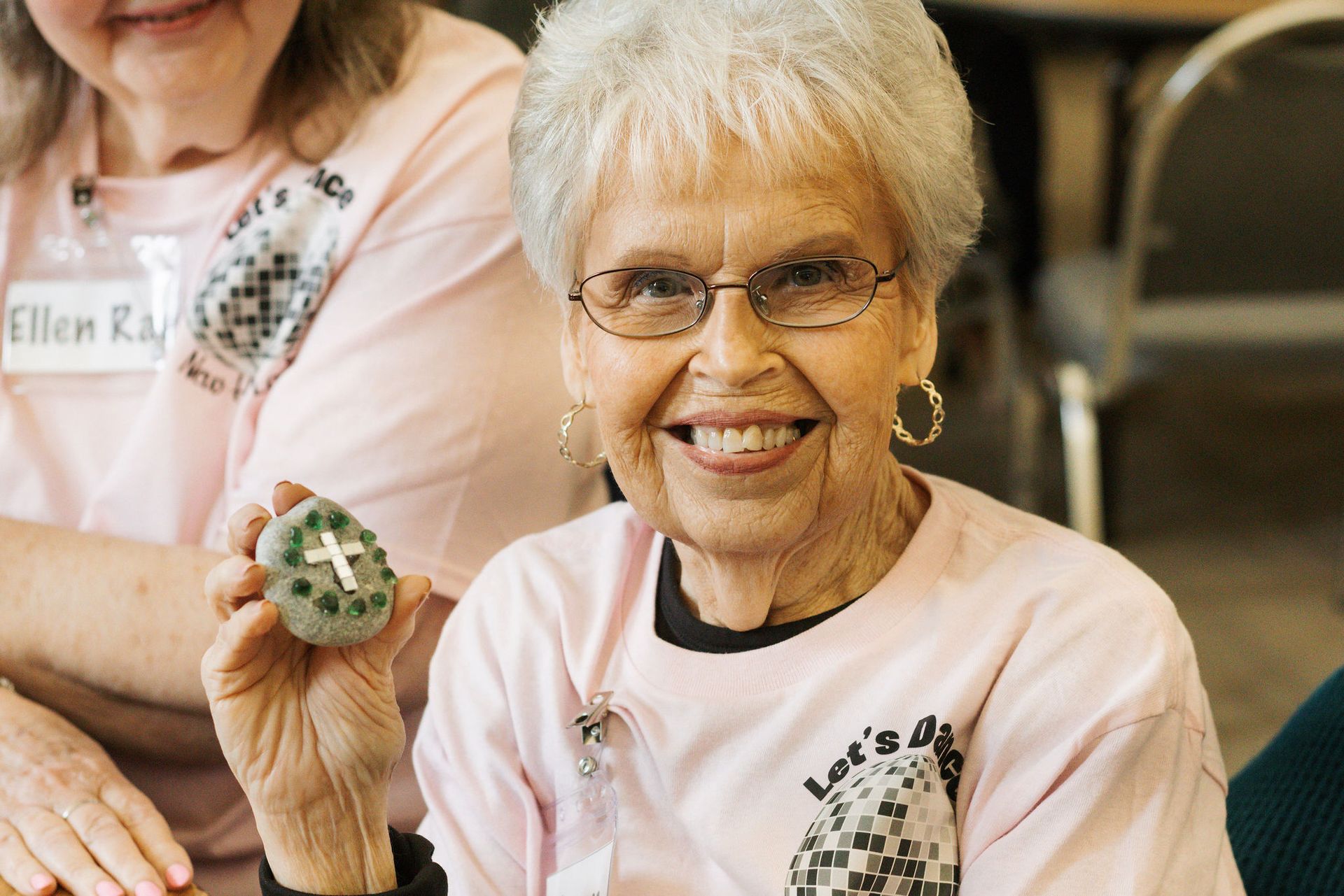 Smiling older woman holding a decorated stone, wearing glasses and a pink shirt.